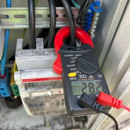 Flow Electric - Miernik cęgowy DT399A pokazuje wartość 28.2 na tle rozdzielnicy elektrycznej z przewodami i złączkami.