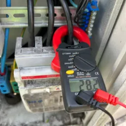 Miernik cęgowy DT399A pokazuje wartość 28.2 na tle rozdzielnicy elektrycznej z przewodami i złączkami.