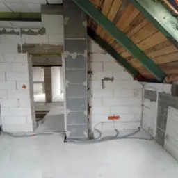 Surowy stan poddasza w trakcie prac instalacyjnych: widoczne przewody elektryczne w szarych peszlach prowadzone po podłodze i ścianach z białych bloczków, pomarańczowe puszki pod gniazdka...