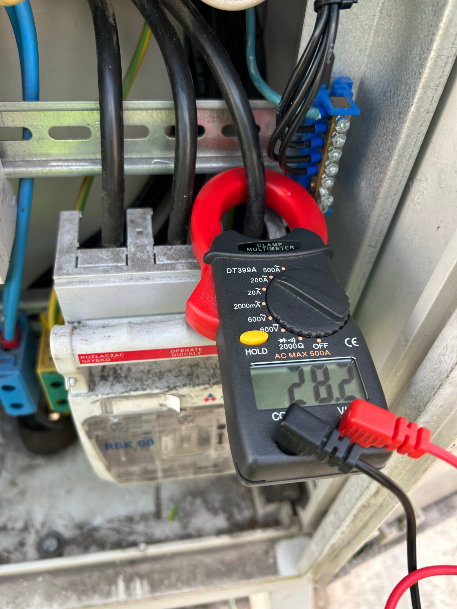 Miernik cęgowy DT399A pokazuje wartość 28.2 na tle rozdzielnicy elektrycznej z przewodami i złączkami.