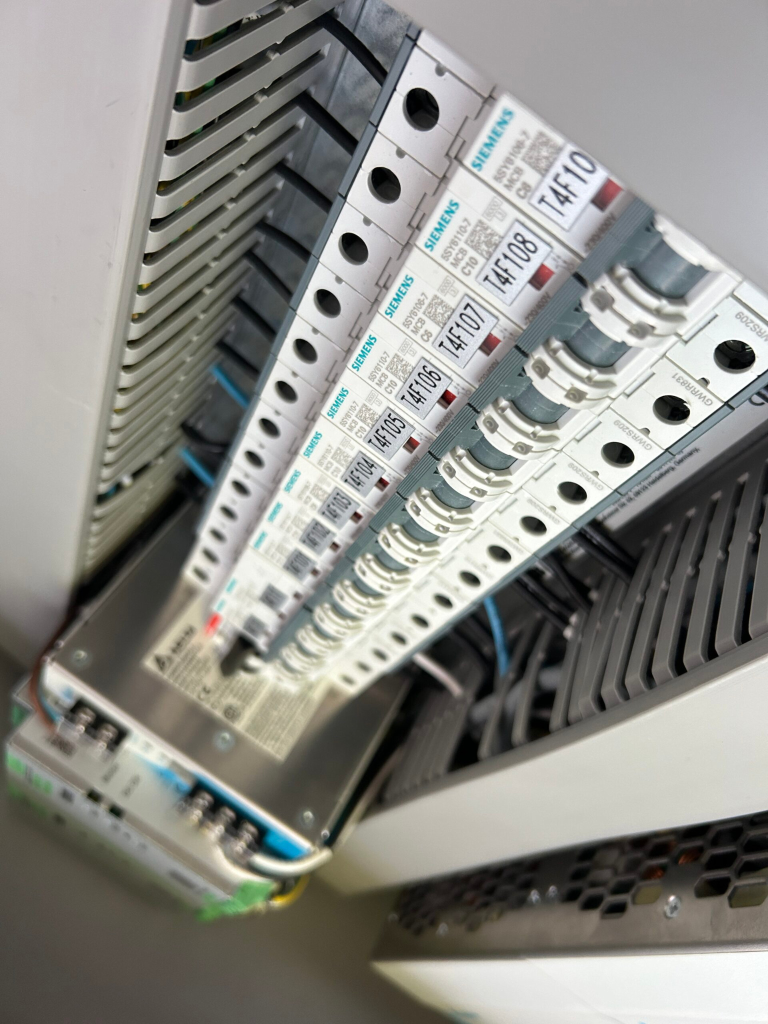 Szczegółowe ujęcie rozdzielnicy elektrycznej z widocznymi bezpiecznikami marki Siemens, ułożonymi w rzędzie, z oznaczeniami T4F103, T4F104, T4F105, T4F106, T4F107, T4F108 i T4F10, oraz widocznymi...