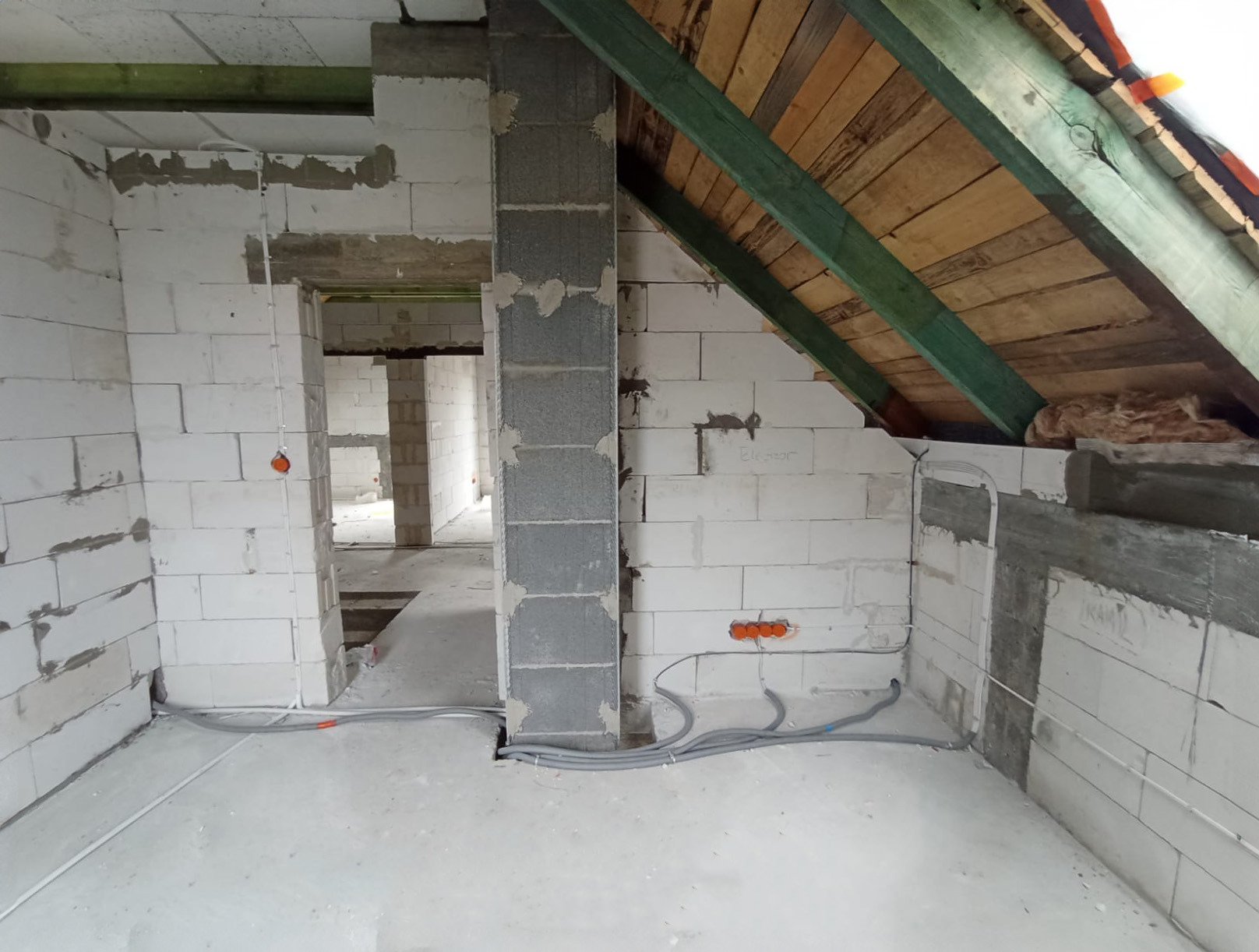 Surowy stan poddasza w trakcie prac instalacyjnych: widoczne przewody elektryczne w szarych peszlach prowadzone po podłodze i ścianach z białych bloczków, pomarańczowe puszki pod gniazdka...