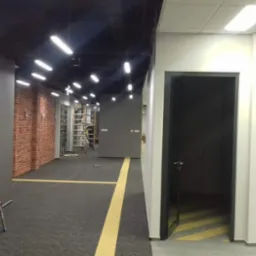 Korytarz biurowy w trakcie remontu z odsłoniętą cegłą, widoczne otwarte drzwi, drabina i instalacja oświetleniowa.