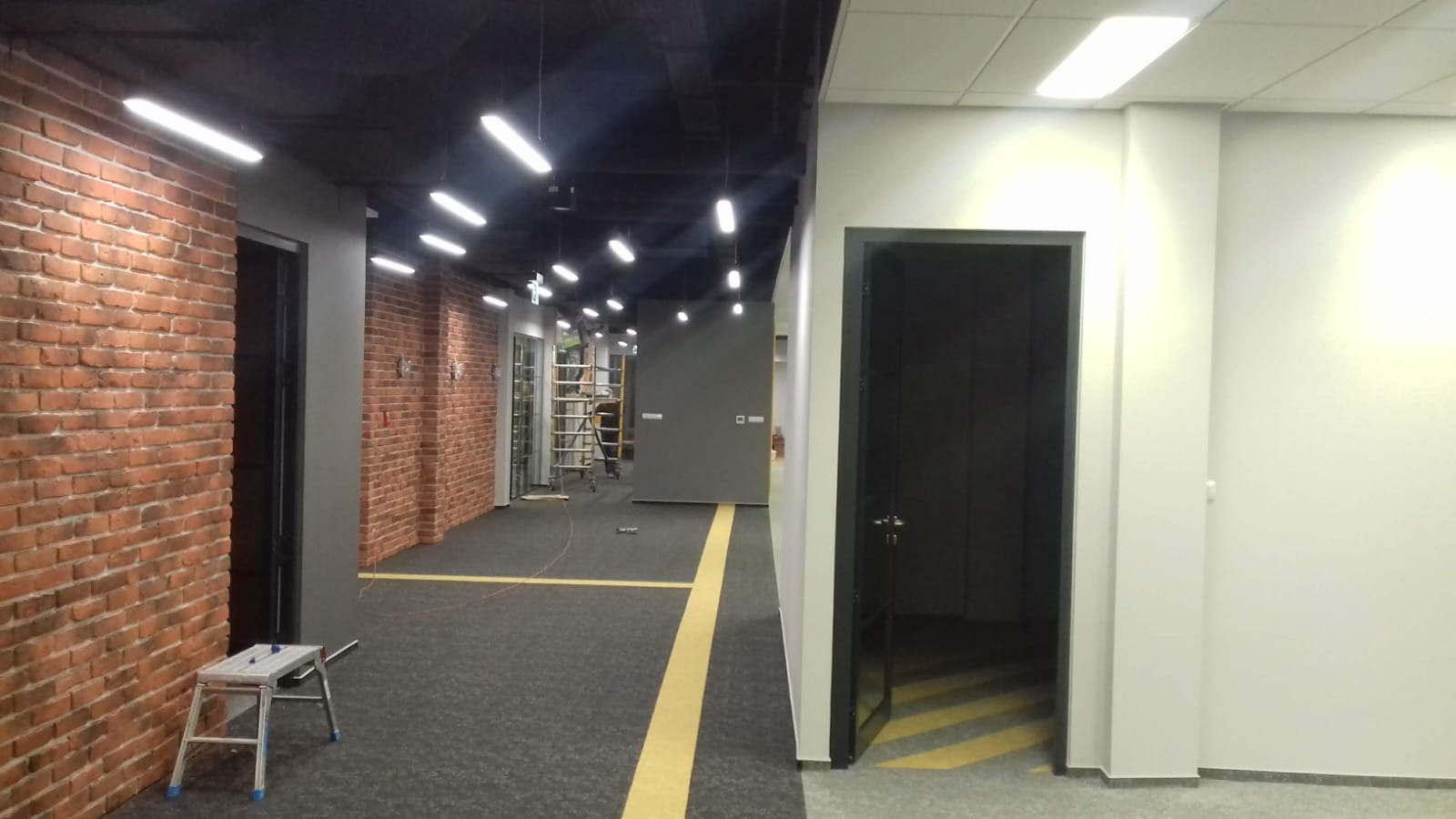 Korytarz biurowy w trakcie remontu z odsłoniętą cegłą, widoczne otwarte drzwi, drabina i instalacja oświetleniowa.