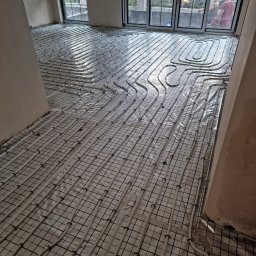 PPHU MUFA - Białe rury ogrzewania podłogowego ułożone na folii i siatce w surowym wnętrzu, widoczne okna z widokiem na zewnątrz i dwie osoby.