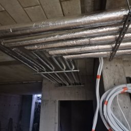 PPHU MUFA - Rury instalacji grzewczej z izolacją aluminiową, zamocowane pod betonowym stropem na metalowych profilach, widoczne także zwinięte kable elektryczne.