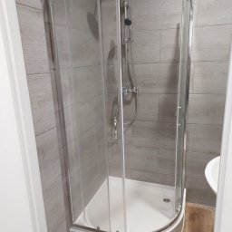 PPHU MUFA - Narożna kabina prysznicowa z chromowanymi profilami i szklanymi drzwiami, zamontowana w łazience z szarymi płytkami imitującymi drewno, widoczna umywalka