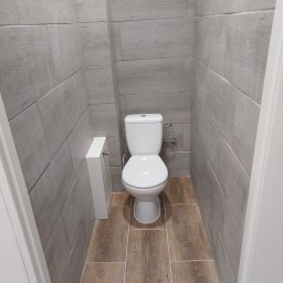 PPHU MUFA - Wąska toaleta wykończona płytkami imitującymi drewno na ścianach i podłodze, widok na sedes i zabudowę geberit.