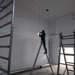 PPHU MUFA - Pracownik na drabinie, używający szlifierki kątowej do cięcia metalowego elementu pod sufitem, z widocznymi iskrami i zieloną linią lasera poziomującego na ścianie w tle, w surowym wnętrzu...