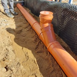 PPHU MUFA - Pomarańczowe rury kanalizacyjne ułożone w wykopie z piasku, widoczna czarna folia izolacyjna na ścianie wykopu i fragmenty sylwetek osób w tle.