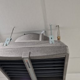 PPHU MUFA - Szary nagrzewnica sufitowa marki Sonniger, zamontowana na metalowych wspornikach, z widocznym podłączeniem hydraulicznym w postaci elastycznego węża, na tle białego sufitu z oświetleniem rastrowym.