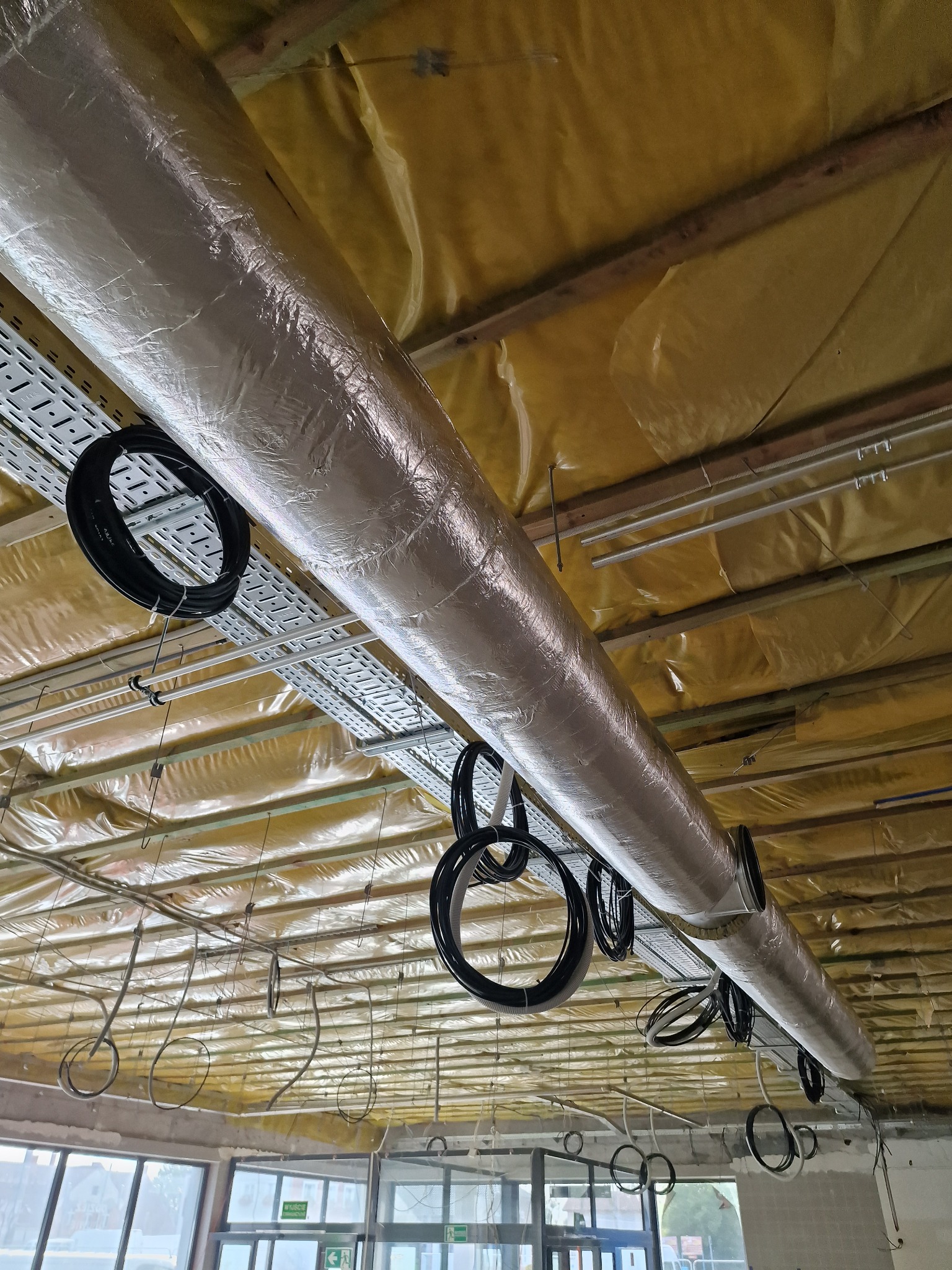 Ocieplony kanał wentylacyjny pod sufitem w trakcie montażu, widoczne kable, metalowe korytka kablowe i żółta folia izolacyjna.