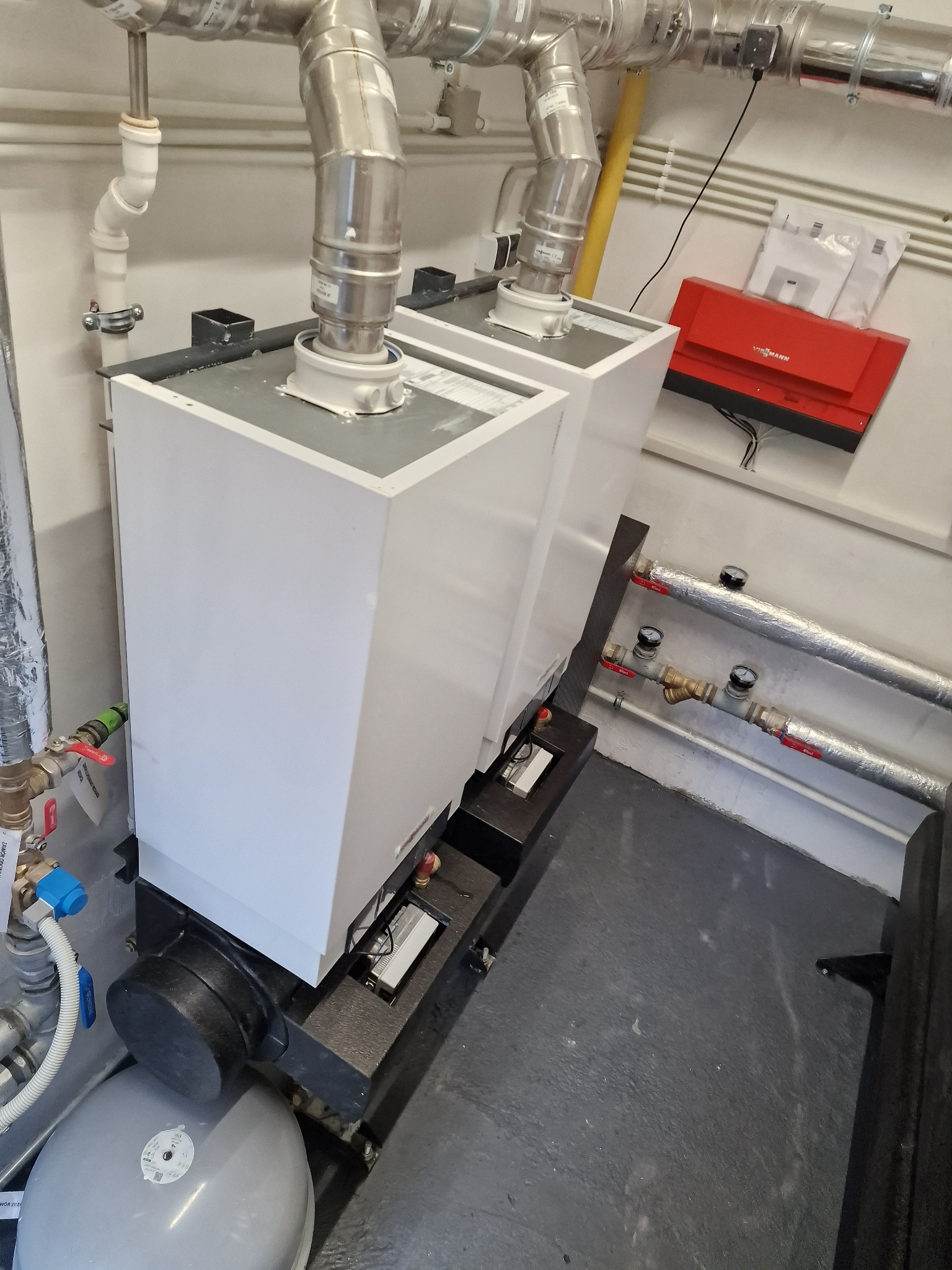 Dwa białe kotły grzewcze z podłączonymi rurami spalinowymi i instalacją hydrauliczną, widoczny czerwony moduł sterujący Viessmann na ścianie.