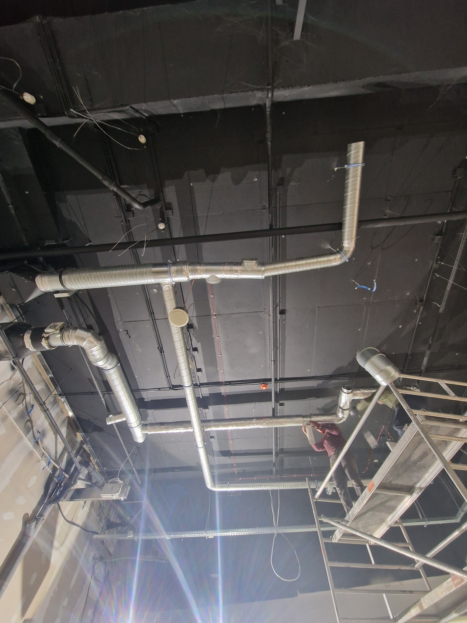 Montaż instalacji wentylacyjnej pod sufitem w Legnicy, widoczny pracownik na rusztowaniu i fragment sufitu z przewodami.