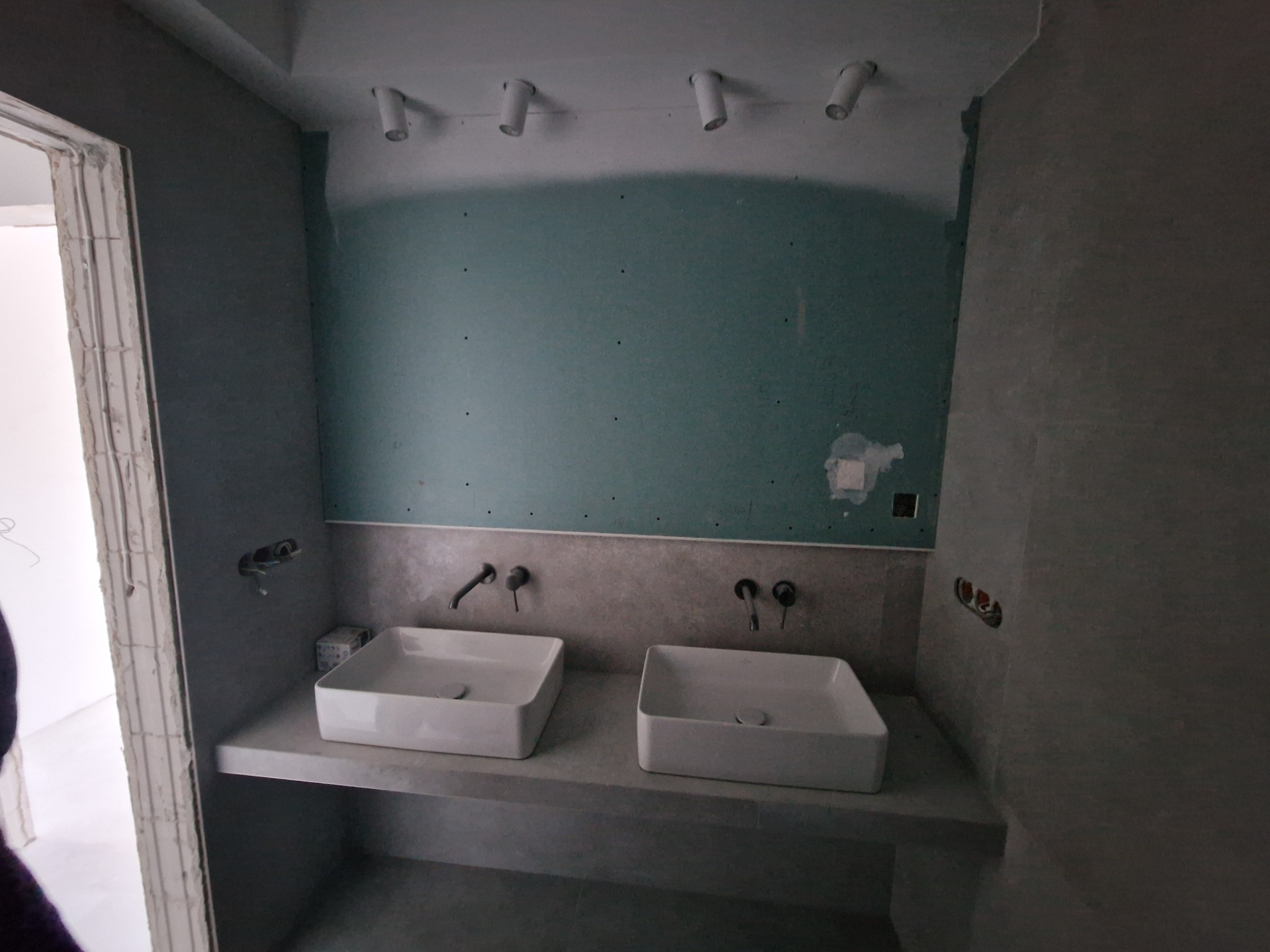 Łazienka w trakcie remontu: dwie białe, kwadratowe umywalki nablatowe z zamontowanymi bateriami na tle ściany z płyt kartonowo-gipsowych, widoczne punkty oświetleniowe na suficie i niedokończone...