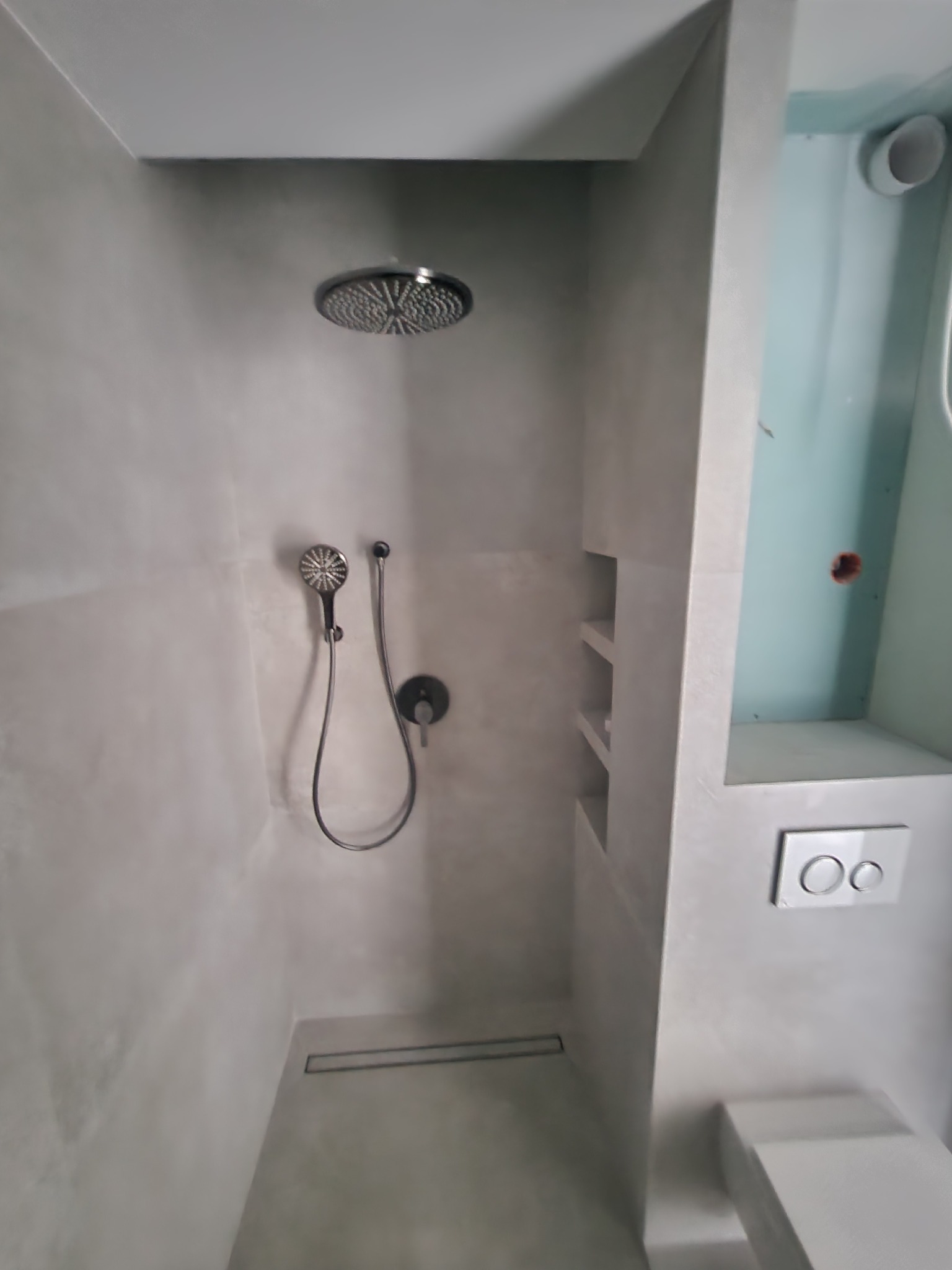 Nowoczesna kabina prysznicowa z deszczownicą i odpływem liniowym w podłodze, wykończona szarym tynkiem dekoracyjnym, z wnęką na kosmetyki i podtynkowym zestawem WC.