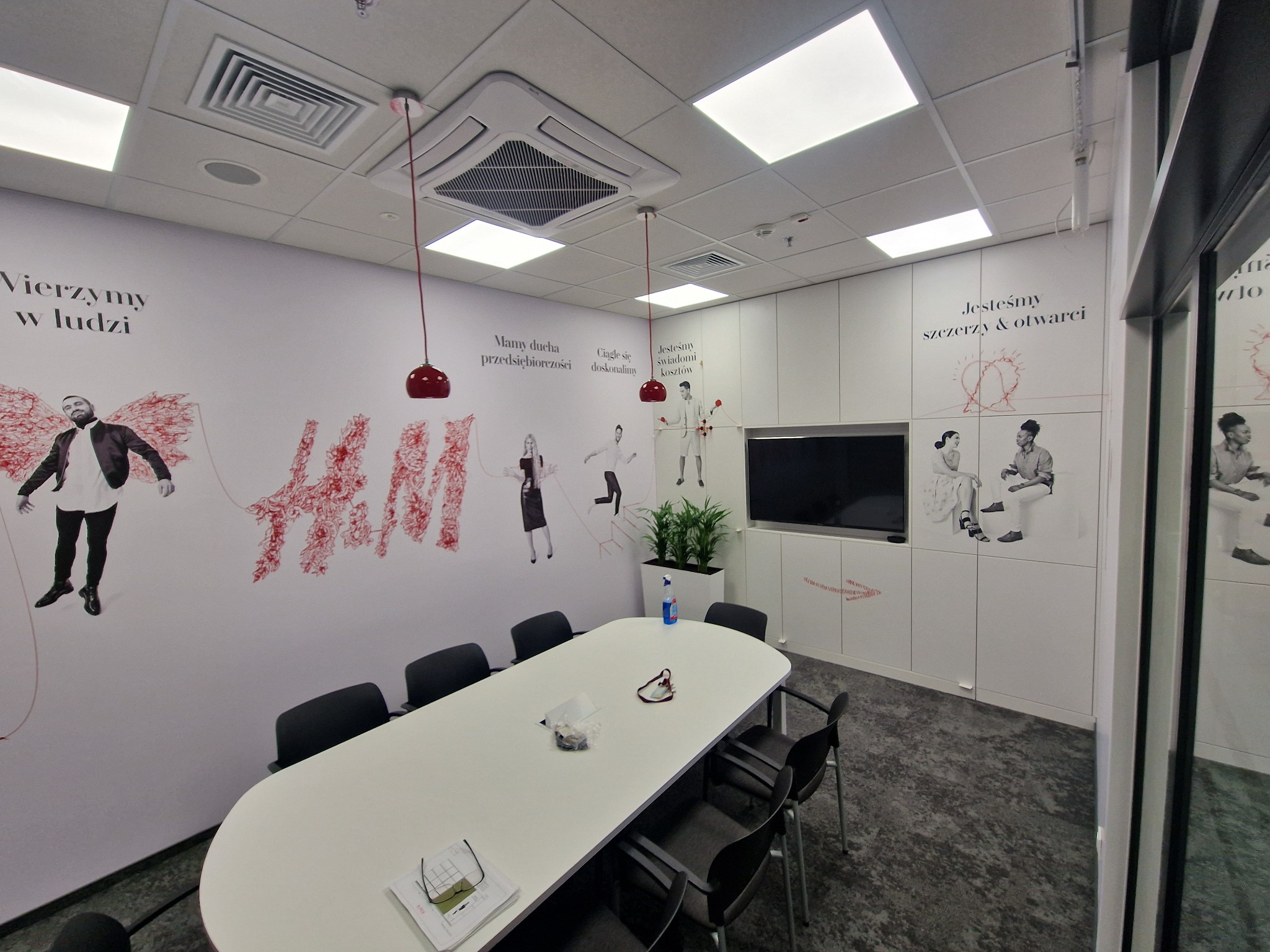 Jasne biuro z podłużnym stołem konferencyjnym, czarnymi krzesłami, ścianami z inspirującymi napisami i grafikami w odcieniach czerwieni, oraz wiszącymi lampami z czerwonymi kloszami.