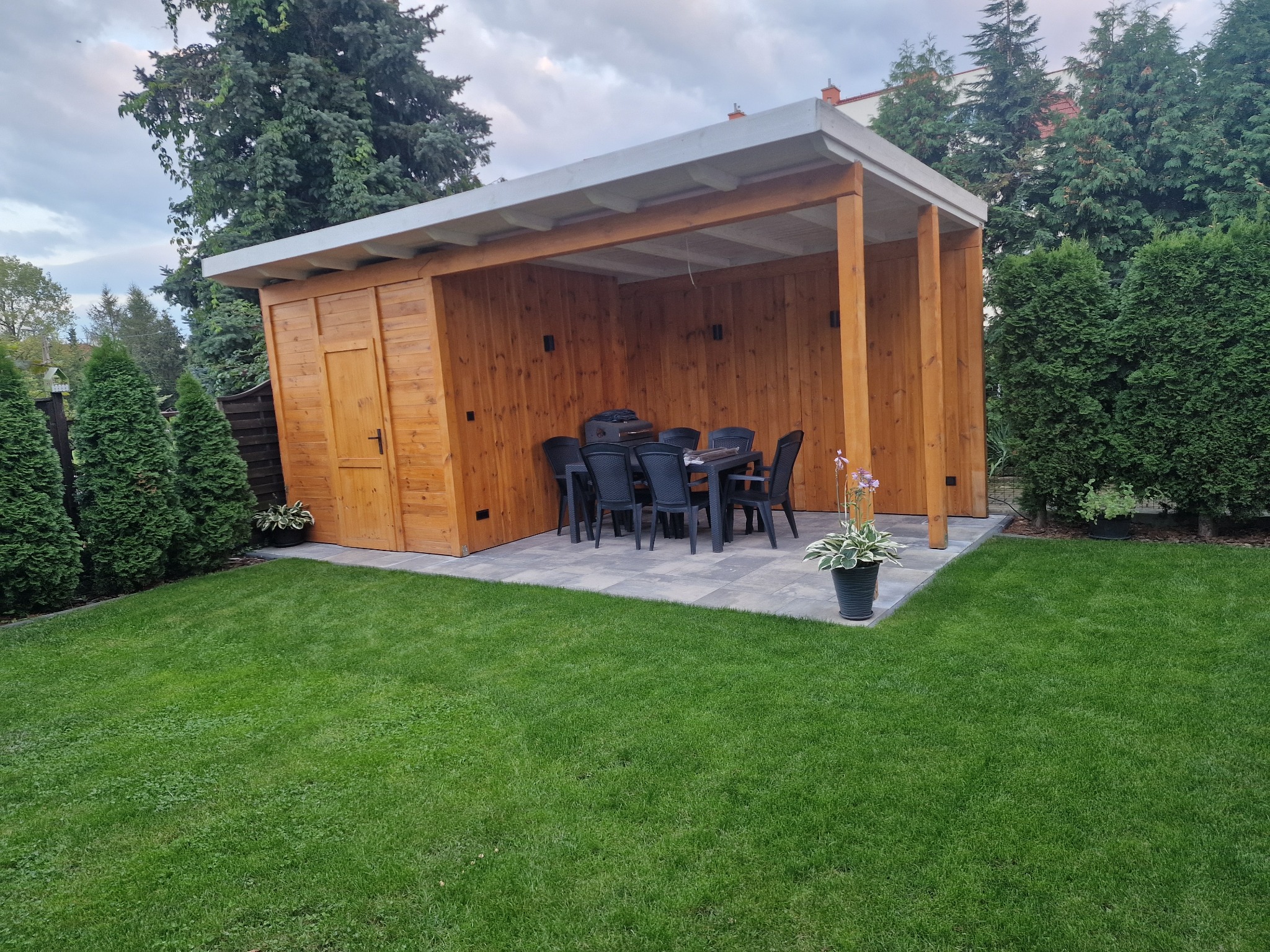 Drewniana altana z przyległym pomieszczeniem gospodarczym, wyposażona w stół i krzesła, ustawiona na wybrukowanym tarasie w zadbanym ogrodzie.