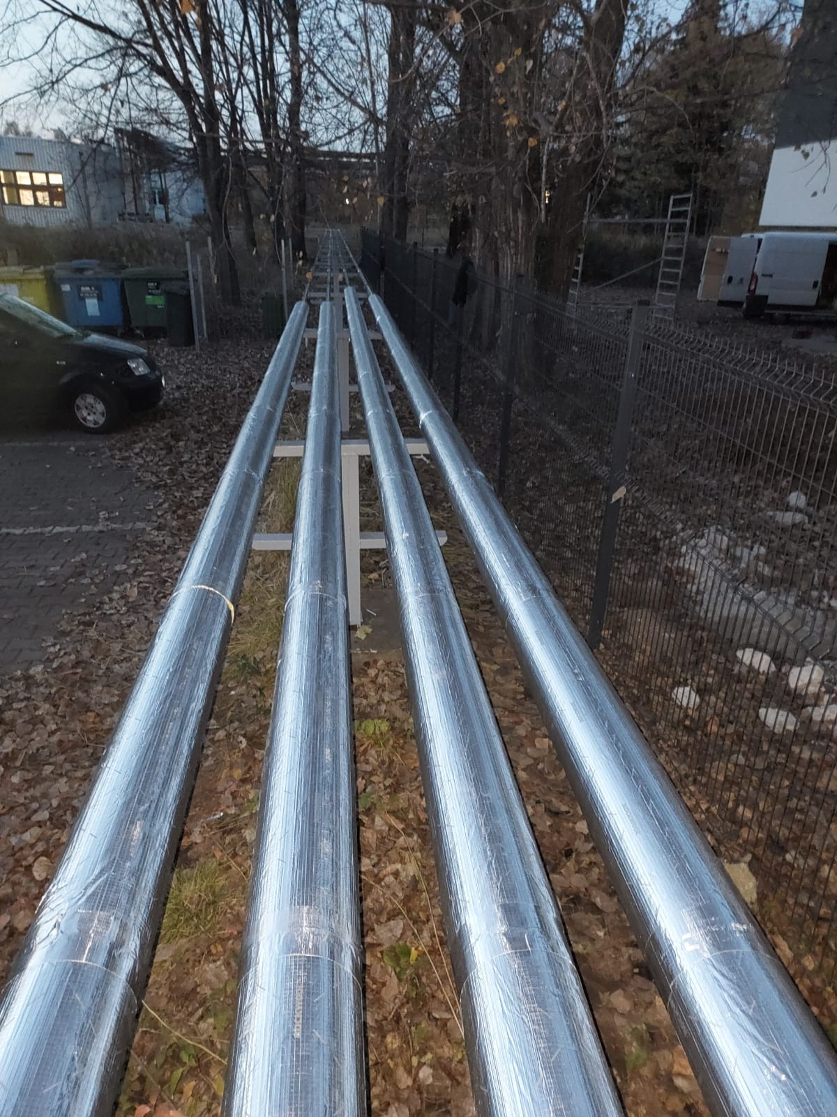 Cztery długie, srebrne rury izolowane termicznie, ułożone równolegle na metalowych podporach na zewnątrz budynku, z widocznymi liśćmi na ziemi i samochodem w tle.