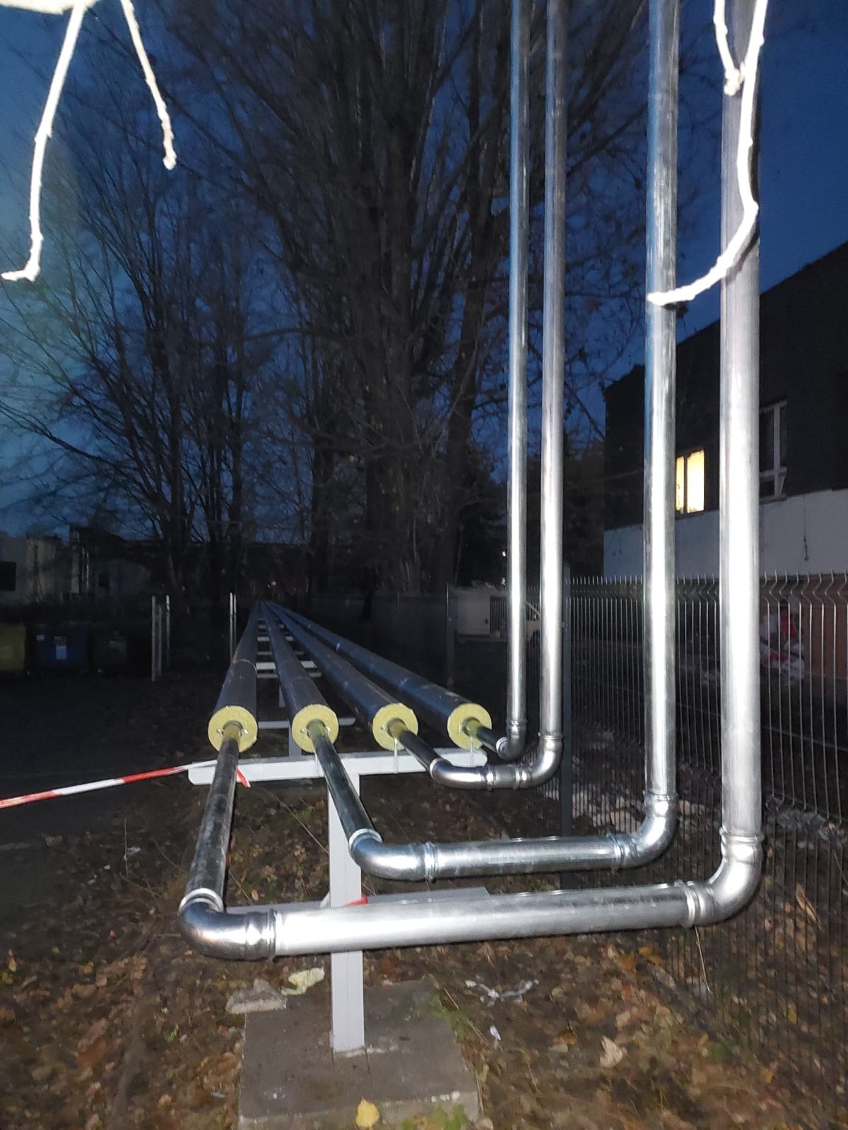 Zewnętrzna instalacja rurociągowa z izolacją termiczną, rury stalowe prowadzone nad ziemią na wspornikach, widoczne zabezpieczenia taśmą ostrzegawczą, w tle drzewa i budynek oświetlony od wewnątrz.