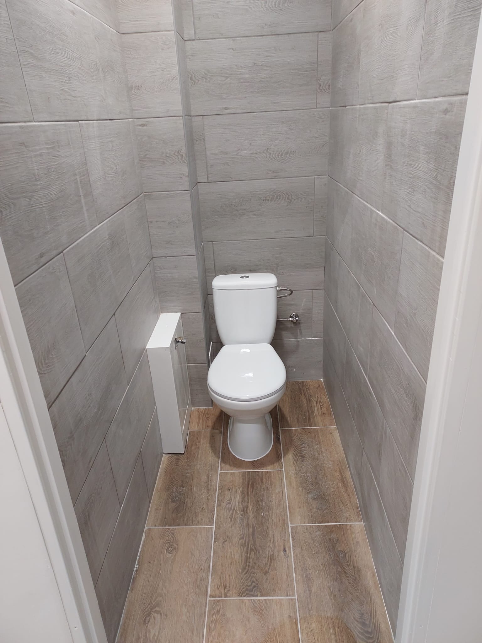 Wąska toaleta wykończona płytkami imitującymi drewno na ścianach i podłodze, widok na sedes i zabudowę geberit.