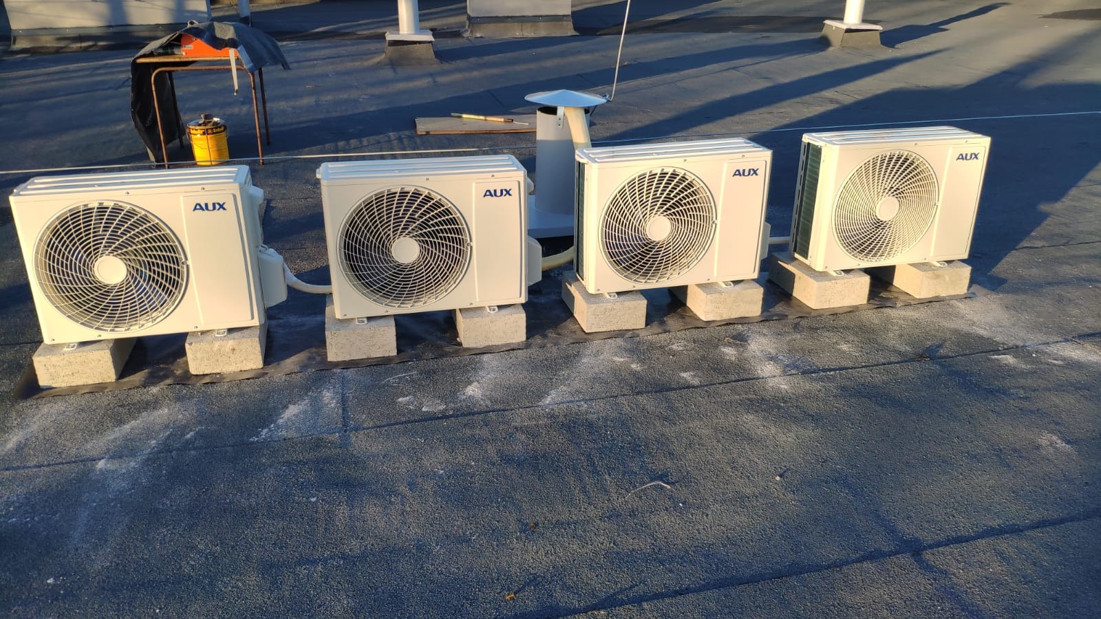 Cztery jednostki zewnętrzne klimatyzacji marki AUX ustawione na betonowych bloczkach na płaskim, bitumicznym dachu, z widocznym osprzętem i narzędziami w tle.