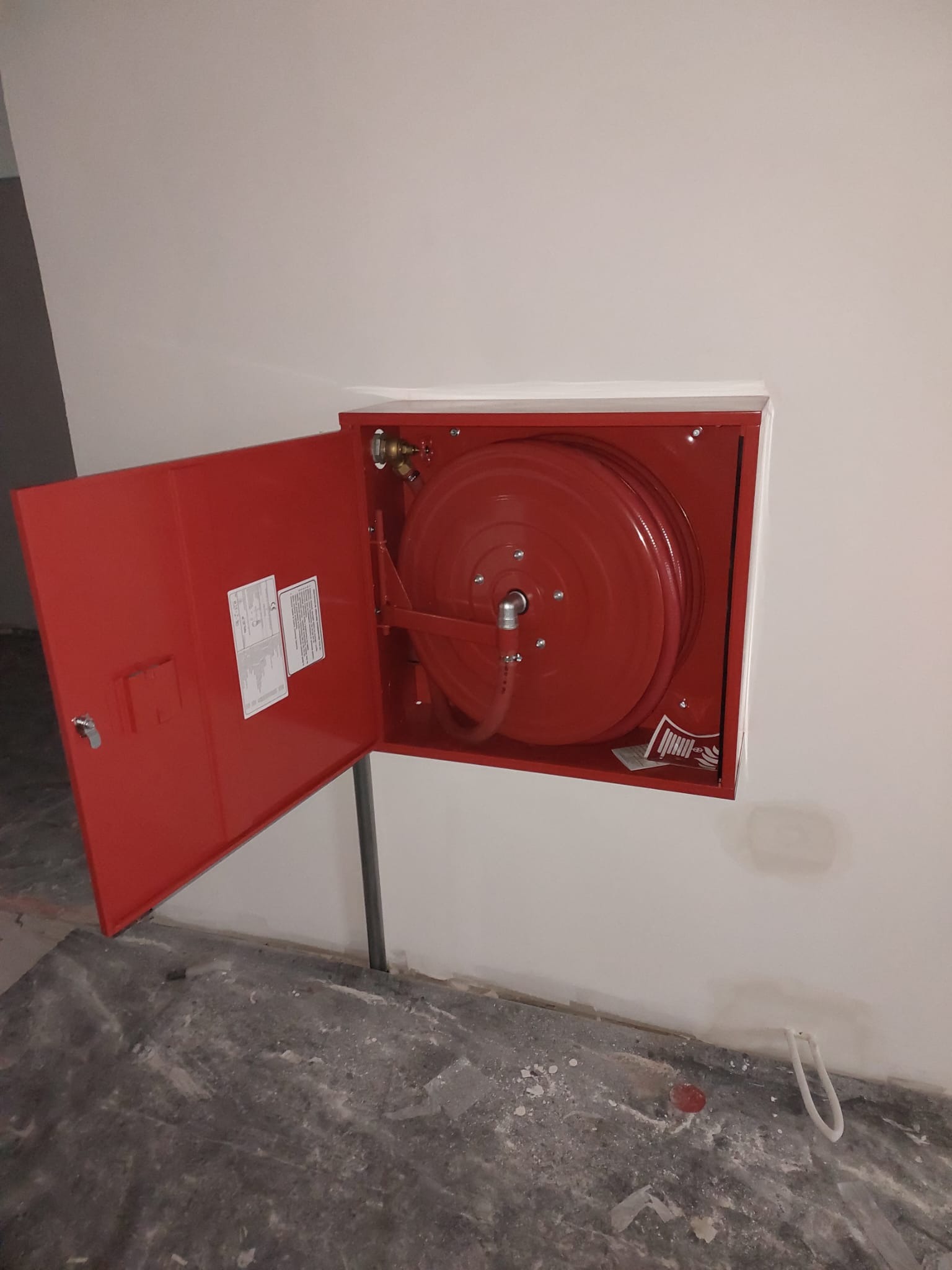 Czerwona szafka hydrantowa z rozwiniętym wężem na bębnie, zamontowana na białej ścianie, z otwartymi drzwiczkami i widoczną dokumentacją techniczną wewnątrz.