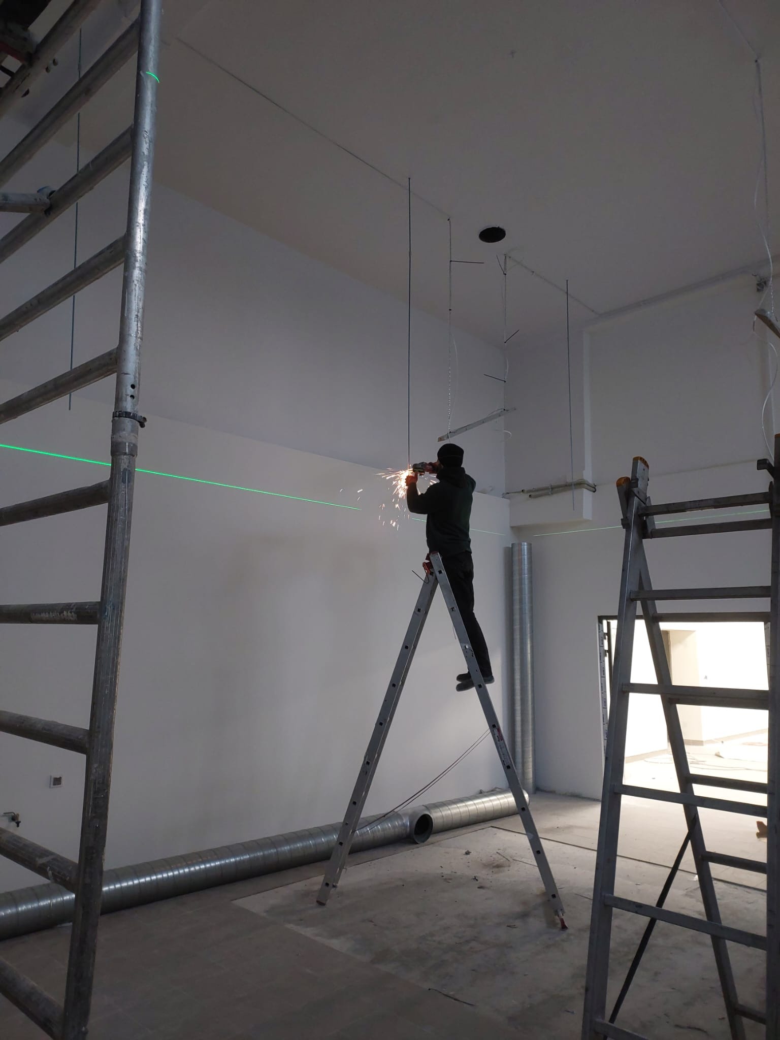 Pracownik na drabinie, używający szlifierki kątowej do cięcia metalowego elementu pod sufitem, z widocznymi iskrami i zieloną linią lasera poziomującego na ścianie w tle, w surowym wnętrzu...
