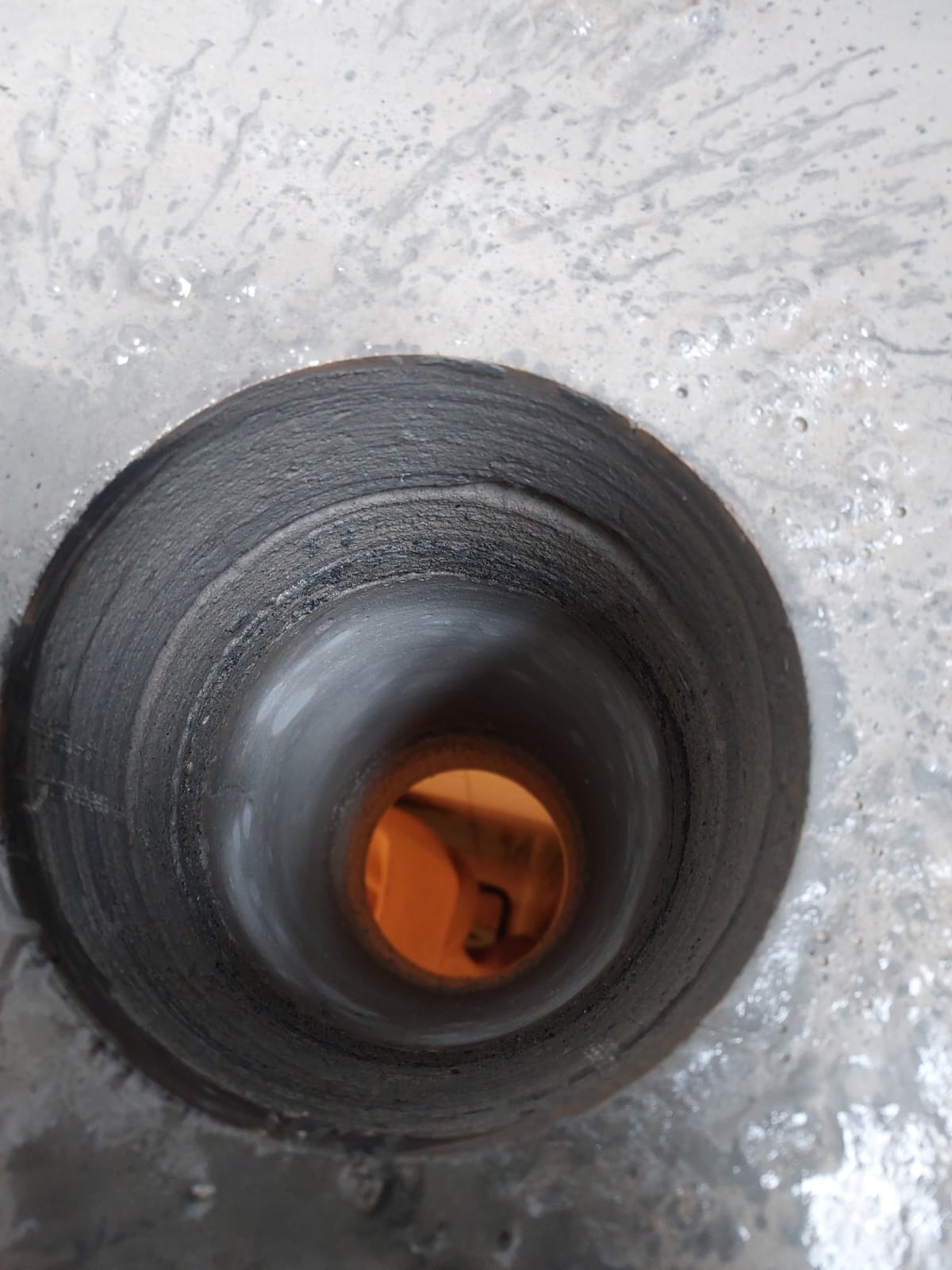 Otwór wywiercony w szarej, betonowej posadzce, widoczne ślady wiercenia i woda, w głębi otworu pomarańczowe światło.