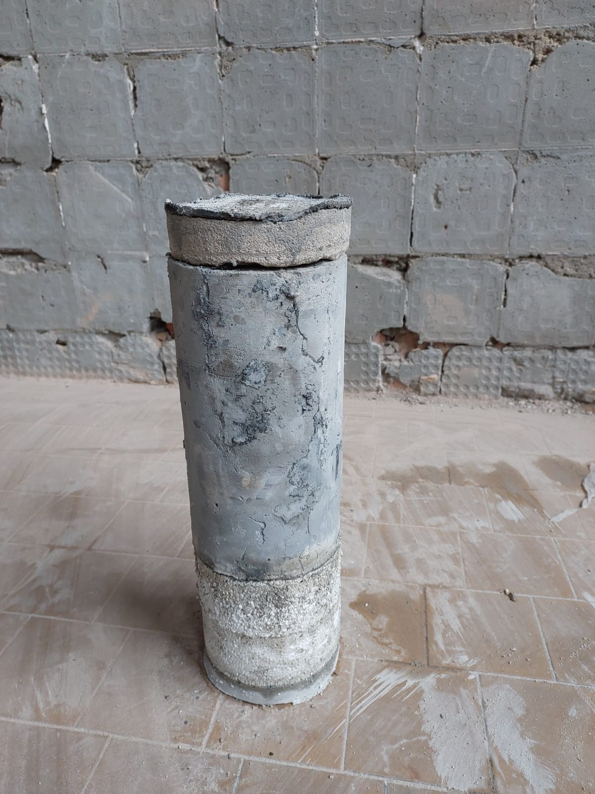 Rdzeń betonowy z widocznymi warstwami i spękaniami, pobrany z podłogi podczas remontu, na tle ściany z odłupanymi płytkami.