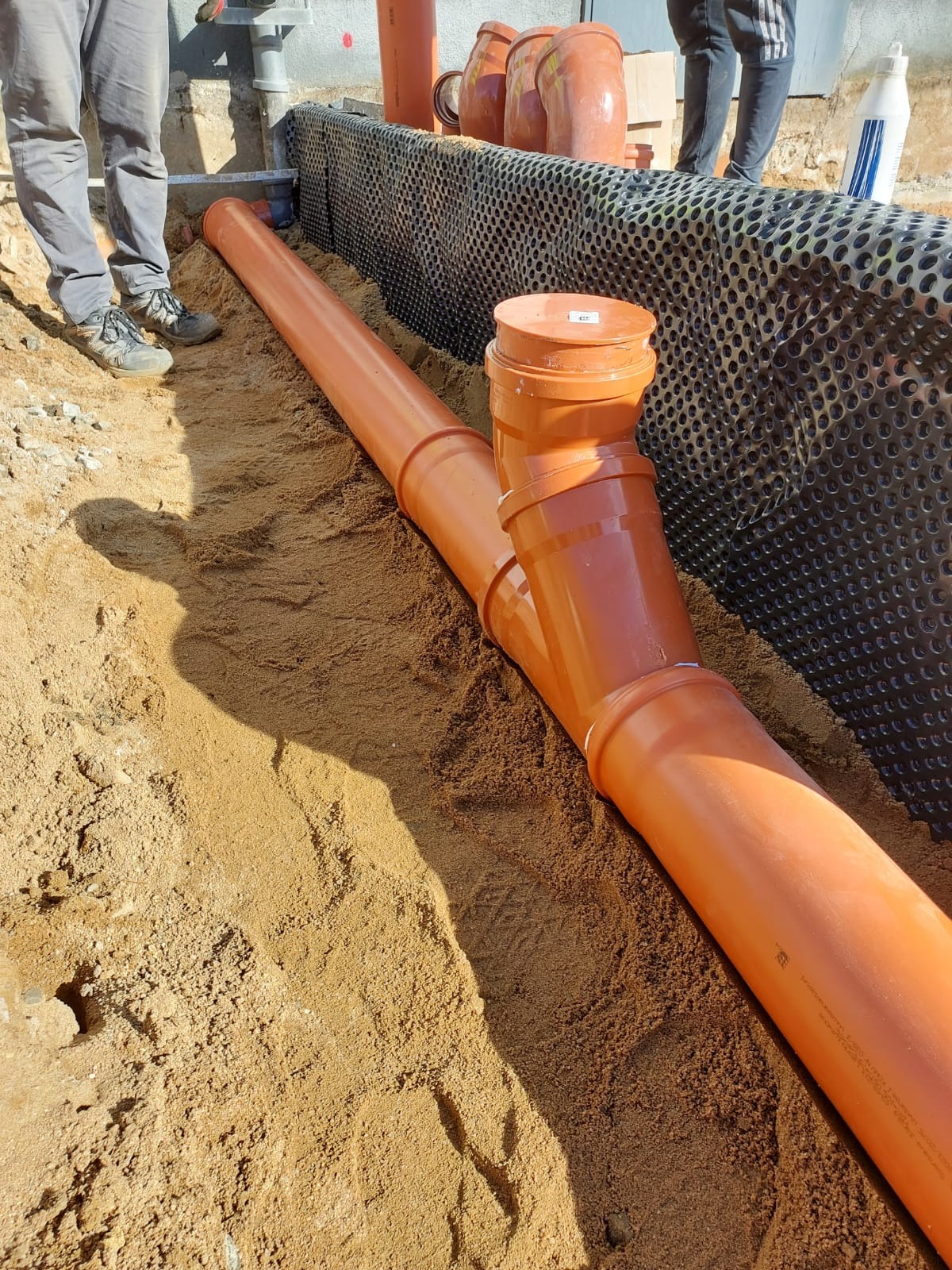Pomarańczowe rury kanalizacyjne ułożone w wykopie z piasku, widoczna czarna folia izolacyjna na ścianie wykopu i fragmenty sylwetek osób w tle.