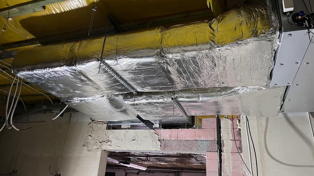 Widok sufitu w trakcie instalacji przewodów wentylacyjnych, owiniętych folią izolacyjną, z widocznymi elementami konstrukcji i przewodami elektrycznymi.