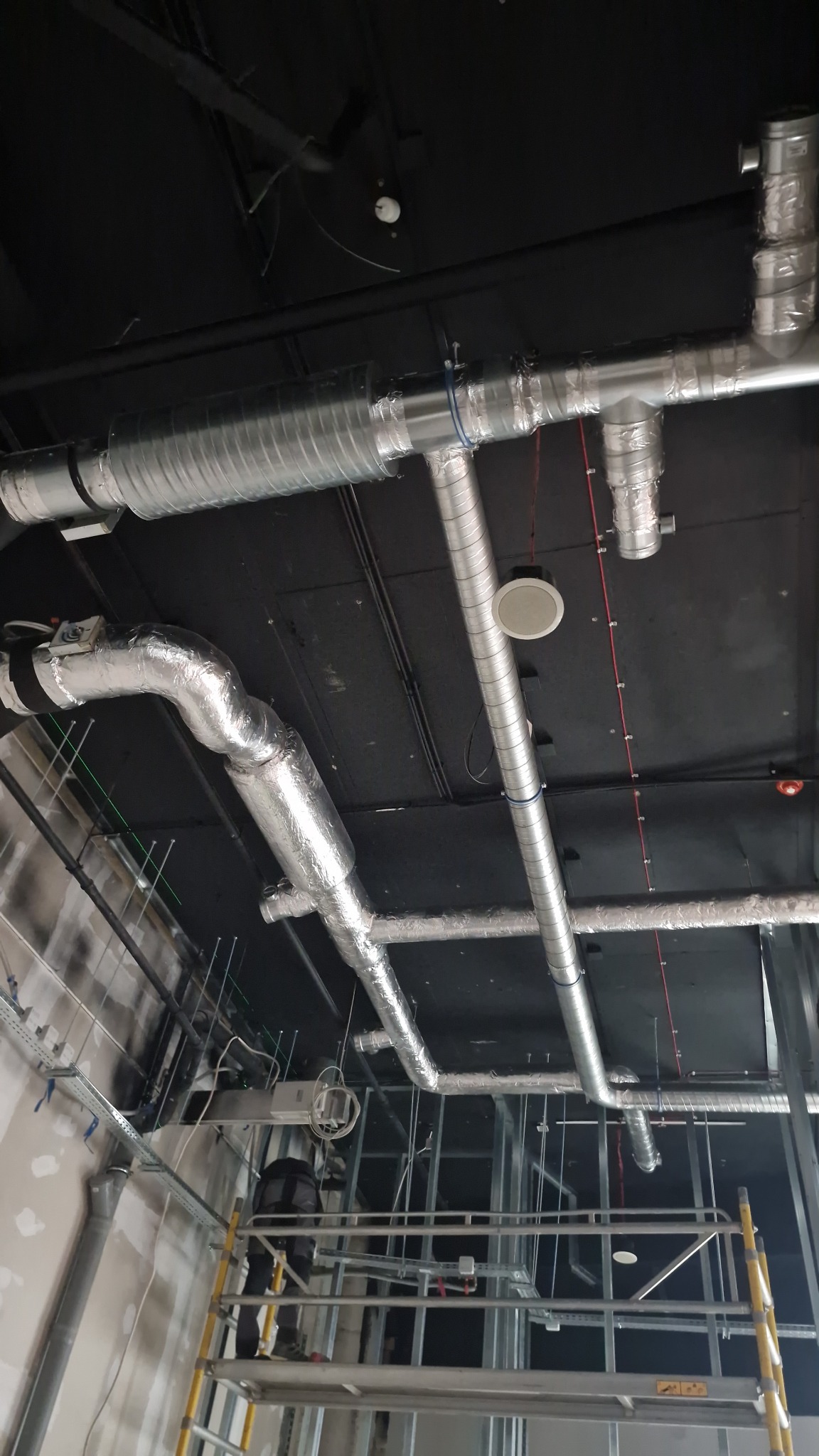 Instalacja wentylacyjna z rur spiro i elastycznych kanałów aluminiowych na czarnym suficie, widoczne elementy rusztowania i okablowania.