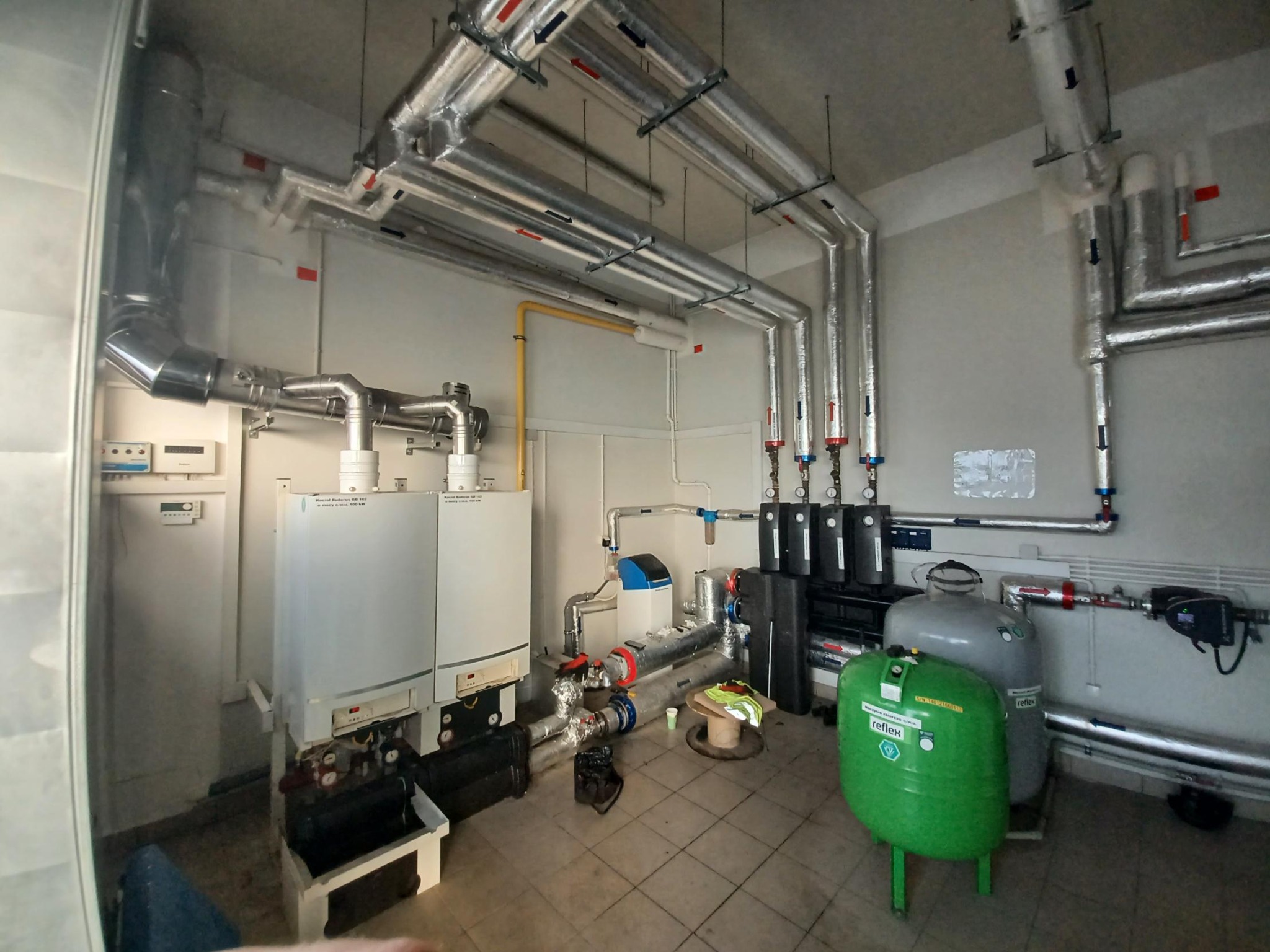 Kompleksowa kotłownia z dwoma kotłami, zbiornikami wyrównawczymi Reflex, licznymi rurami izolowanymi termicznie oraz elementami instalacji hydraulicznymi i elektrycznymi.