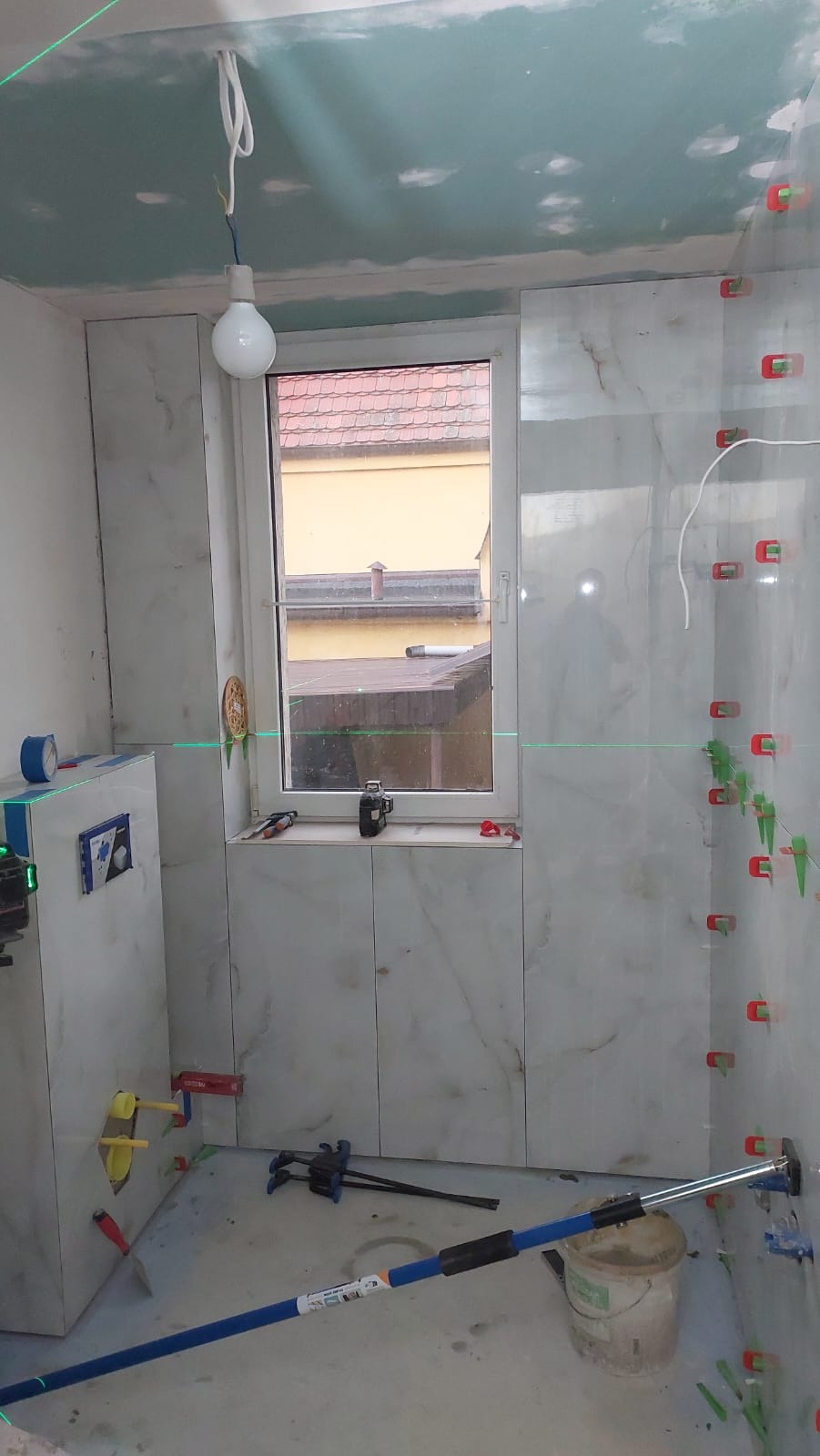 Łazienka w trakcie wykańczania: białe płytki imitujące marmur na ścianach, system poziomowania płytek, laser krzyżowy, surowy sufit z wiszącą żarówką, widok na budynki przez okno.