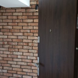 Pełnia - Ciemne, drewnopodobne drzwi wejściowe z metalową klamką, zamontowane obok ściany z dekoracyjnej cegły w odcieniach czerwieni, z czarnym kinkietem i białym sufitem podwieszanym.