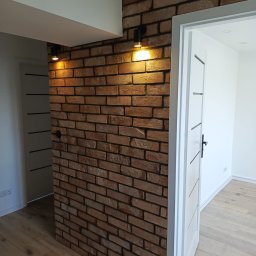 Pełnia - Nowoczesne wnętrze z dekoracyjną ścianą z cegły w ciepłych odcieniach, oświetloną dwoma czarnymi reflektorami. Widoczne białe drzwi i jasne panele podłogowe.