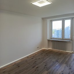 Pełnia - Świeżo wykończony pokój z drewnianymi panelami ułożonymi ukośnie, białymi ścianami, nowoczesną lampą sufitową i dyskretnym oświetleniem LED wzdłuż listew przypodłogowych. Widok z okna na miejski...