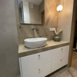 Nowoczesna łazienka z białą szafką pod umywalką, okrągłą umywalką nablatową, lustrem w metalowej ramie i dekoracyjnym oświetleniem.