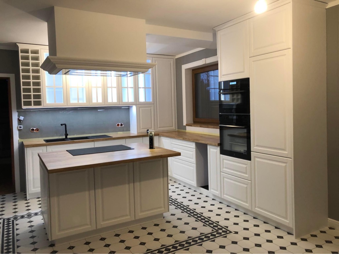 Jasna kuchnia z wyspą, białe szafki z frezowanymi frontami, drewniany blat, czarna armatura i płyta grzewcza, wzorzysta podłoga w romby.