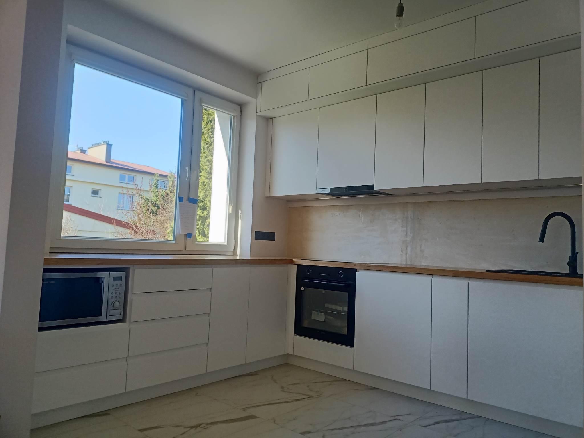 Jasna kuchnia z białymi frontami bez uchwytów, drewnianym blatem i czarną armaturą, widok na budynek przez okno.