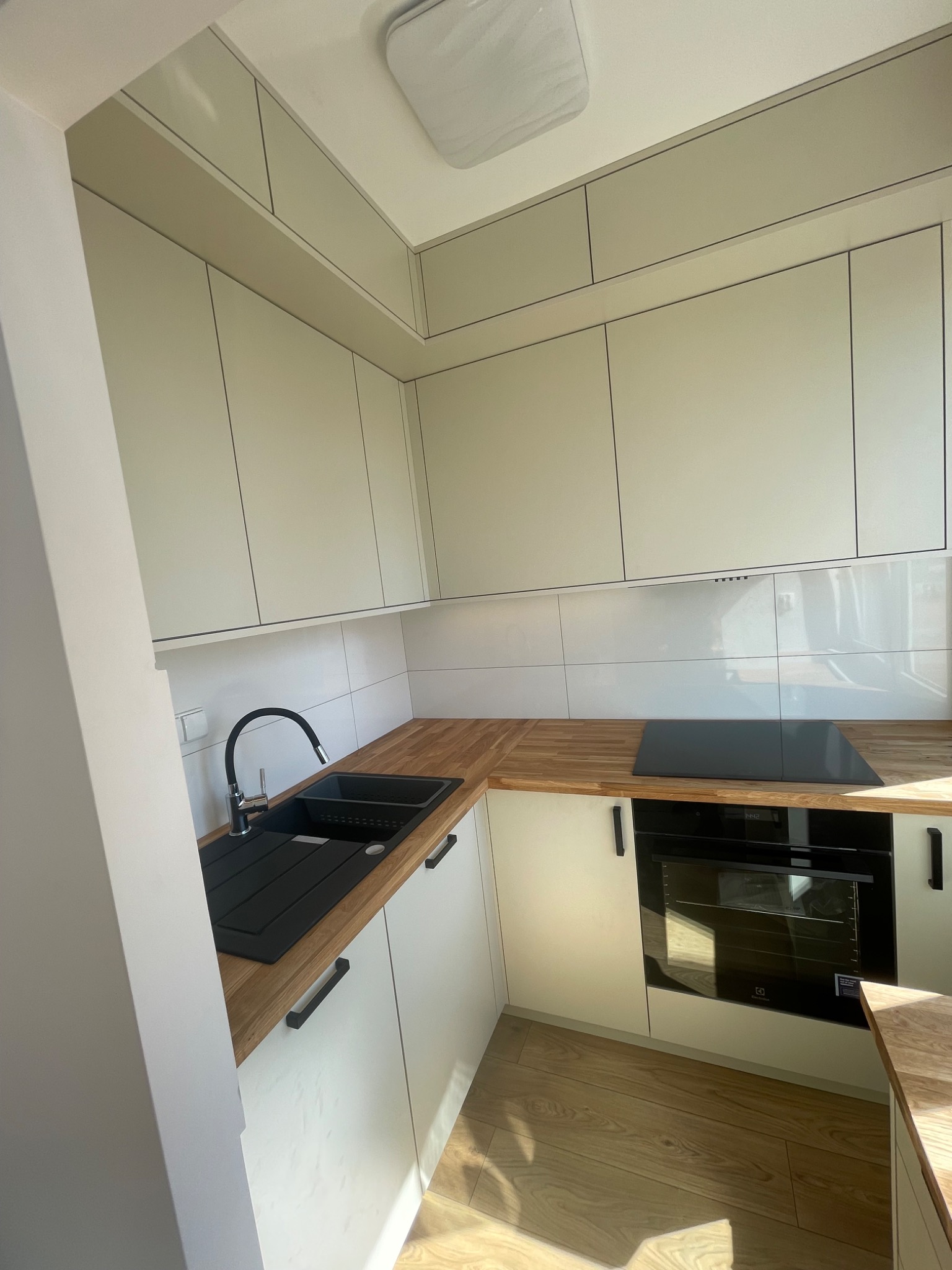 Nowoczesna, jasna kuchnia z białymi szafkami i drewnianym blatem, czarny zlew i bateria, płyta indukcyjna, piekarnik Electrolux.