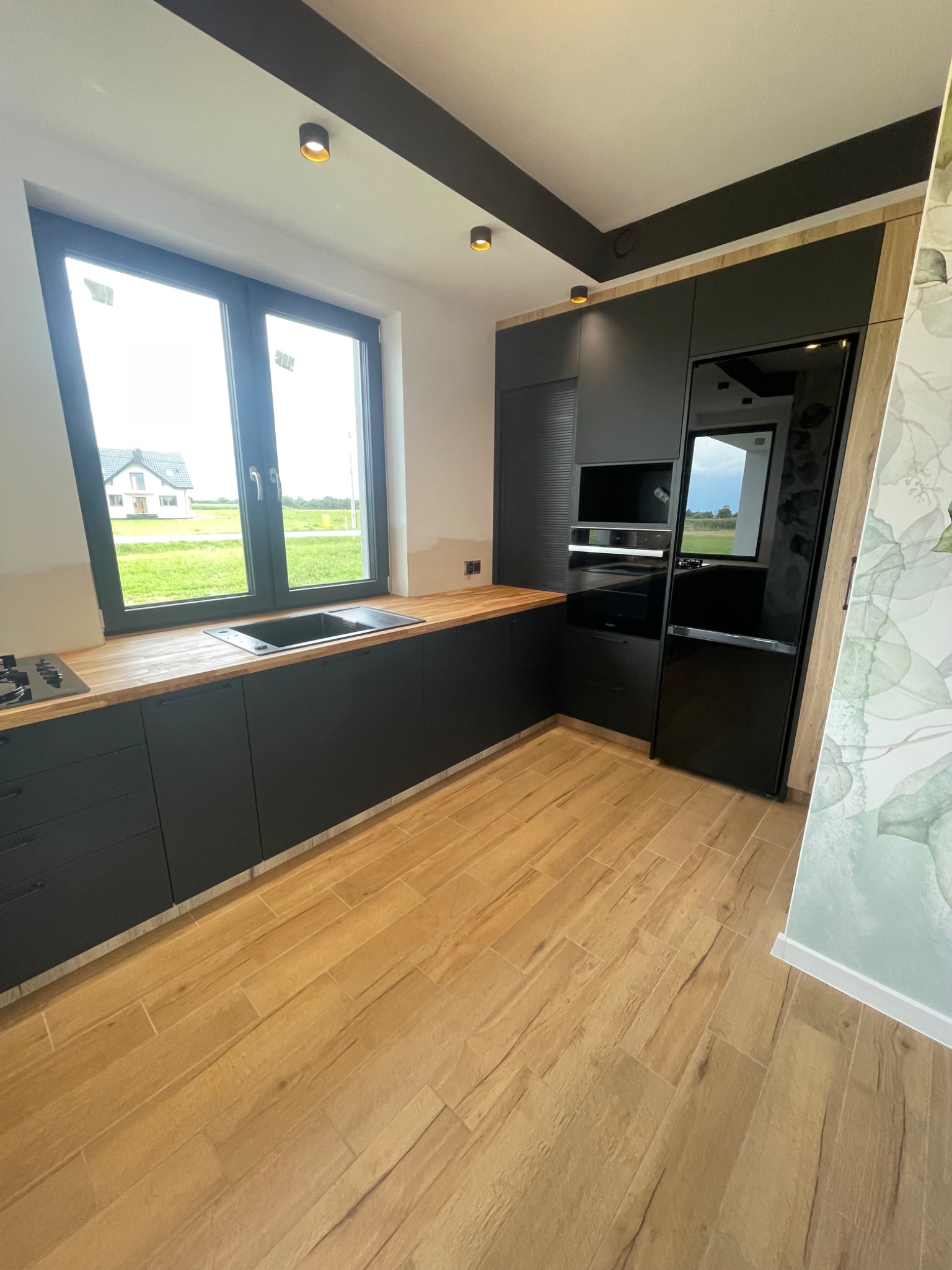 Nowoczesna kuchnia z czarnymi szafkami i drewnianym blatem, widok na okno z zabudową w tle, drewnopodobna podłoga.