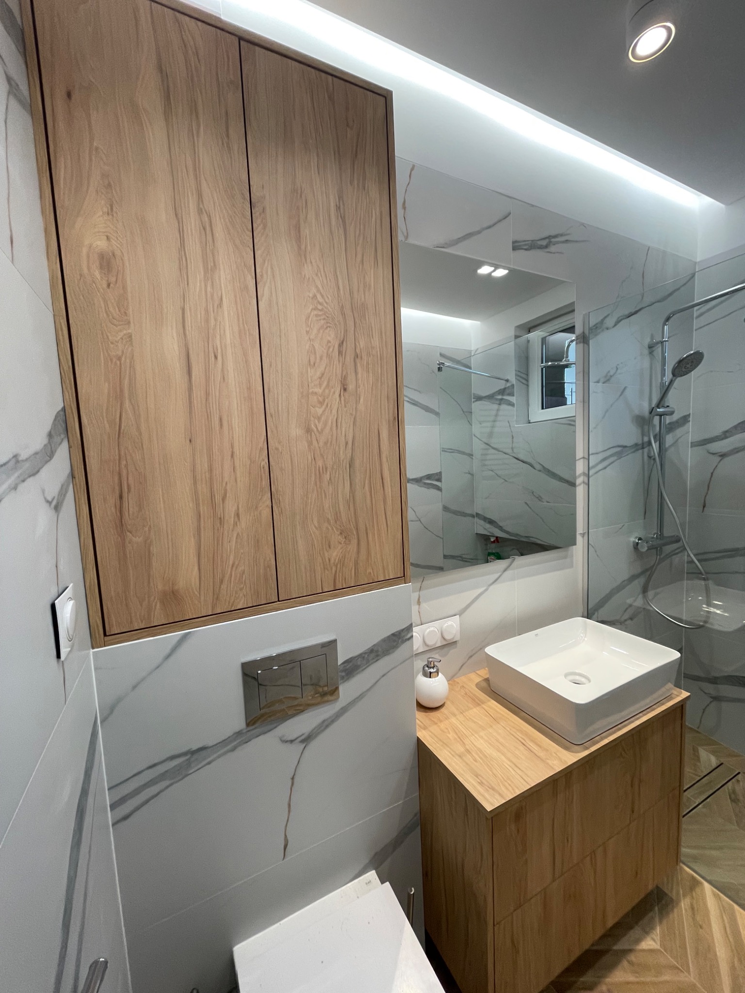 Nowoczesna łazienka z drewnianą szafką pod umywalką, wiszącą szafką z frontami imitującymi drewno, białymi płytkami marmurkowymi i oświetleniem LED.