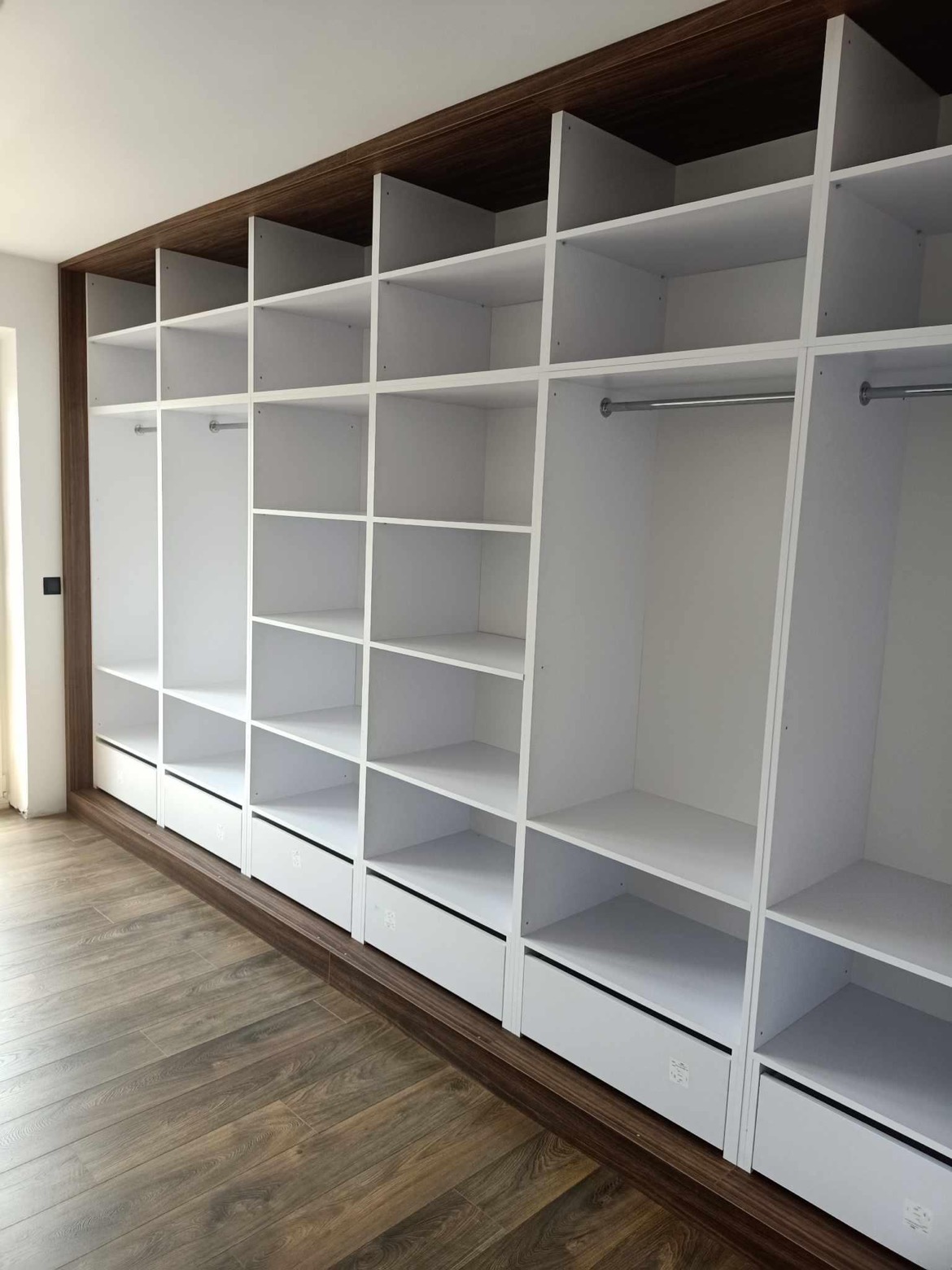 Biała szafa wnękowa z szufladami i półkami, wykończona ciemnym drewnem, widok na drewnianą podłogę.