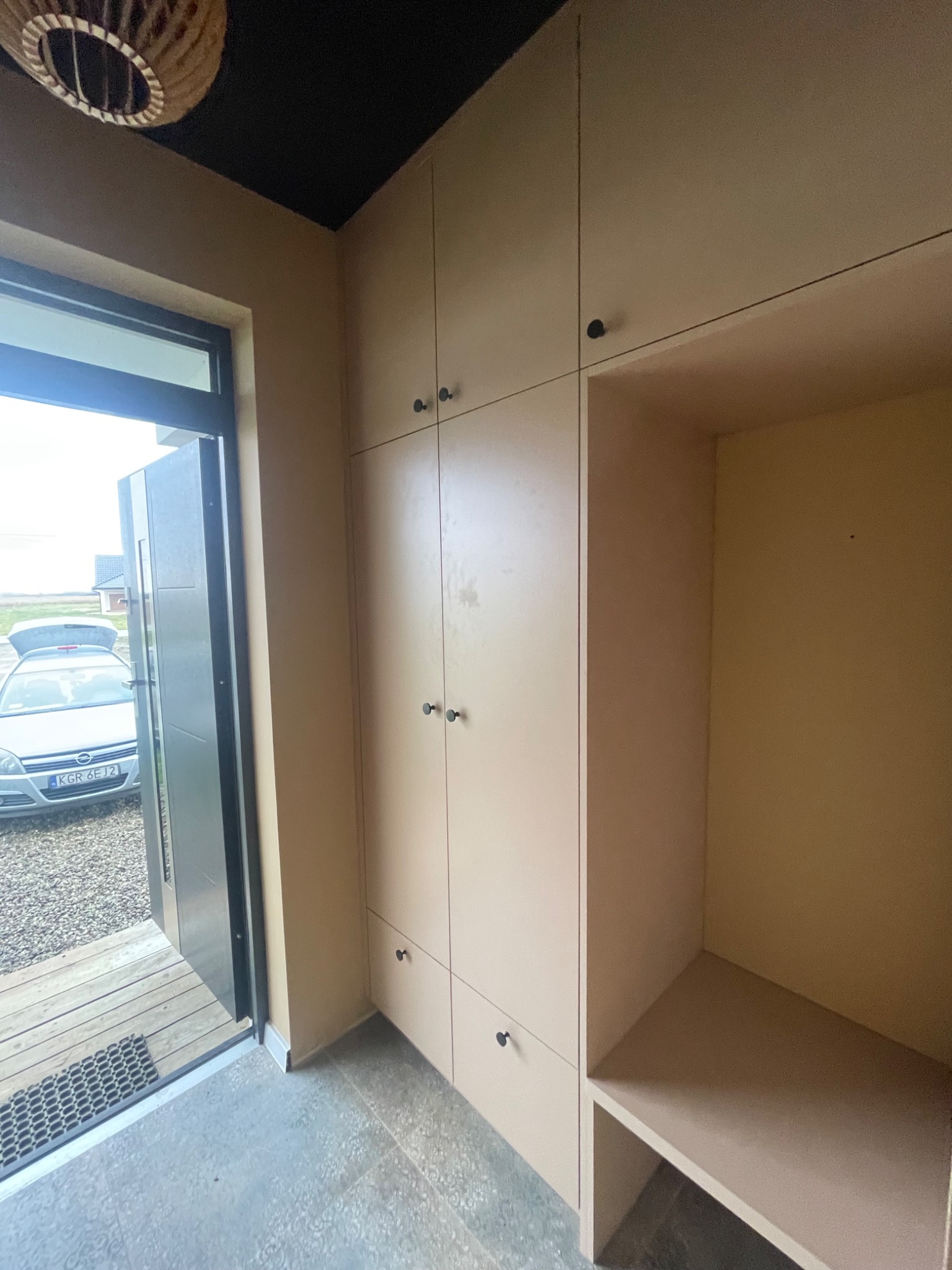 Jasna zabudowa meblowa z szafkami i otwartą wnęką w przedpokoju, widok na otwarte drzwi wejściowe i samochód na zewnątrz.