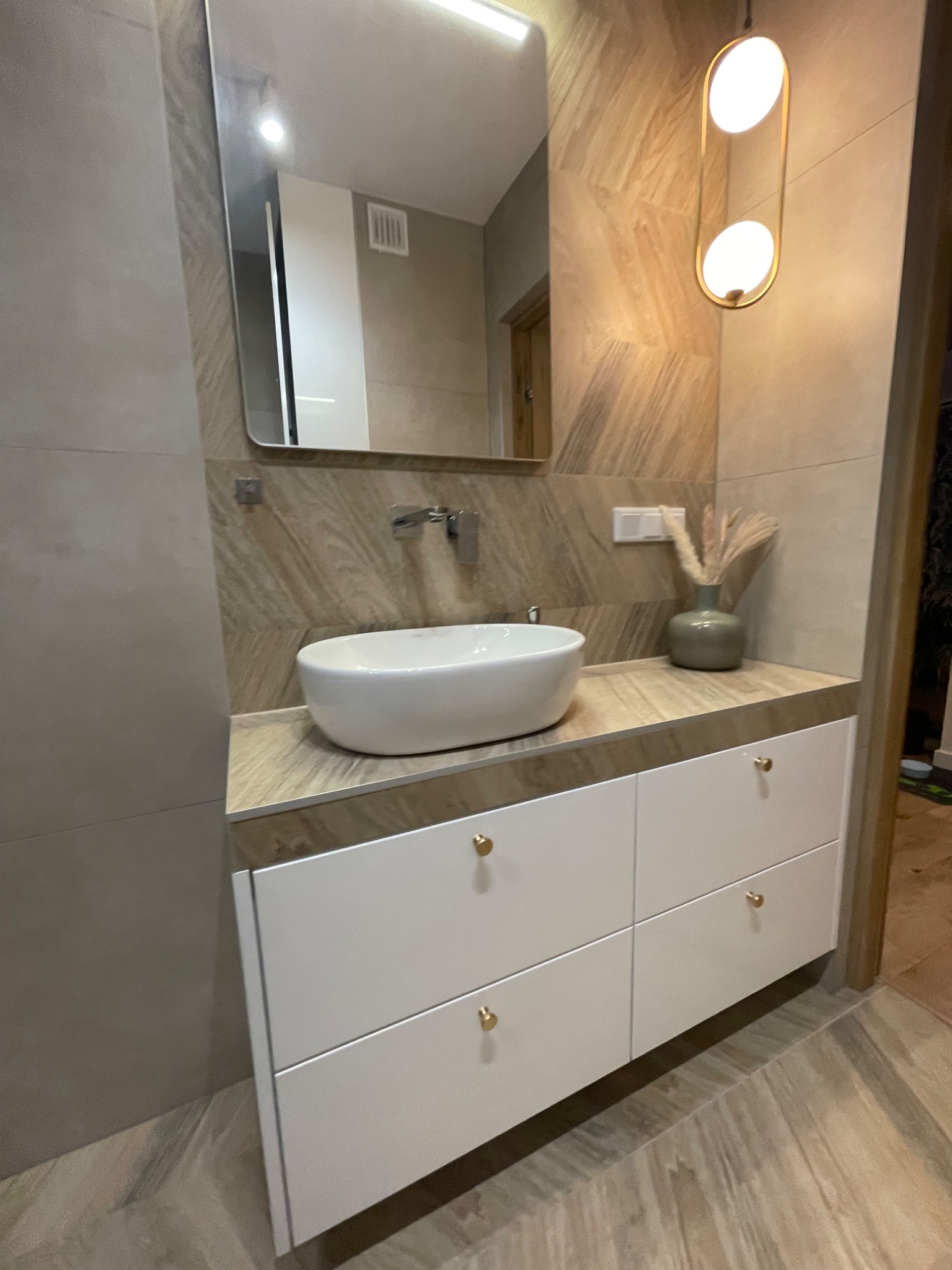 Nowoczesna łazienka z białą szafką pod umywalką, okrągłą umywalką nablatową, lustrem w metalowej ramie i dekoracyjnym oświetleniem.