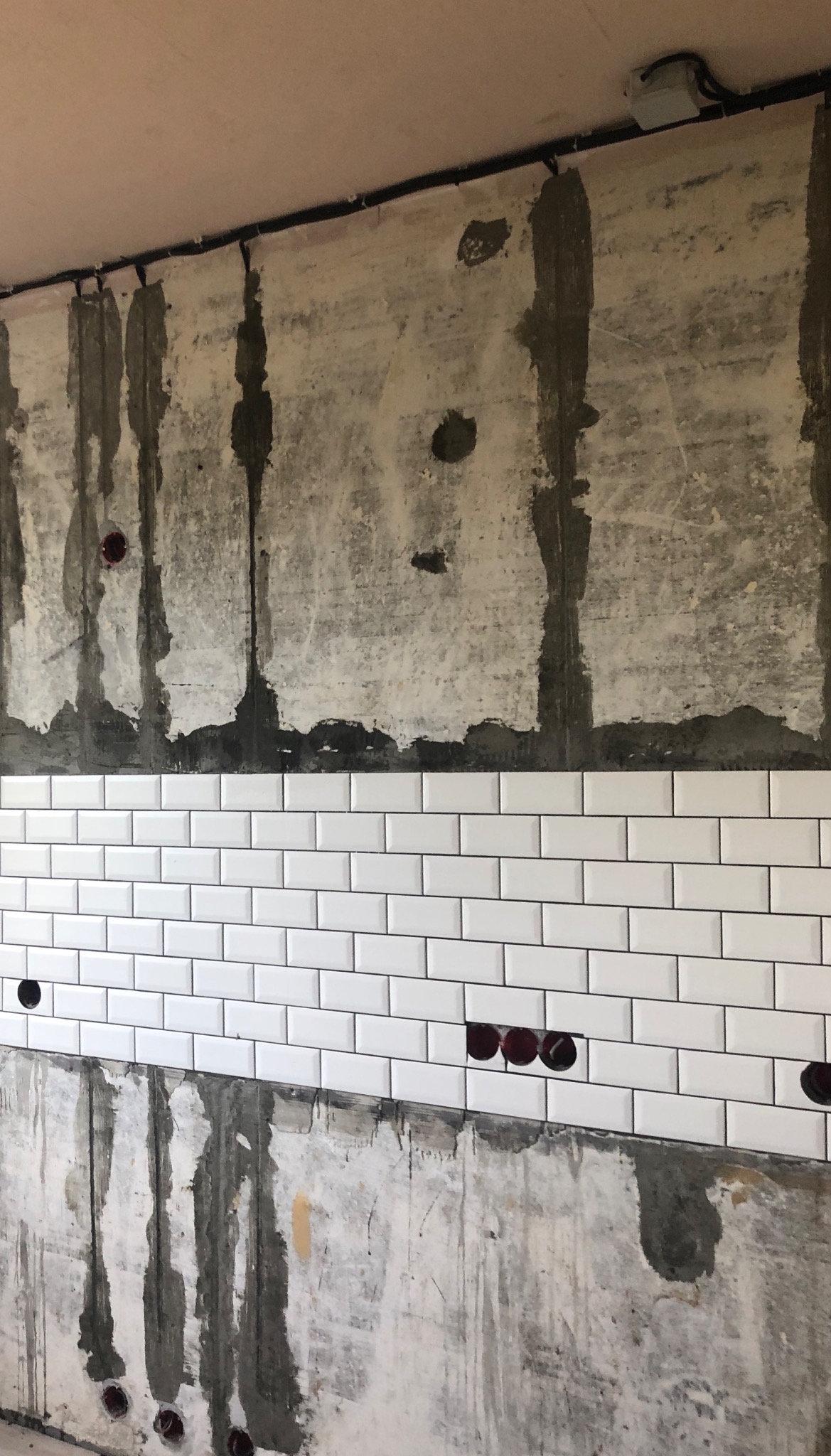 Ściana w trakcie układania białych, prostokątnych płytek, widoczne puszki elektryczne bez osprzętu i surowy beton z pozostałościami zaprawy