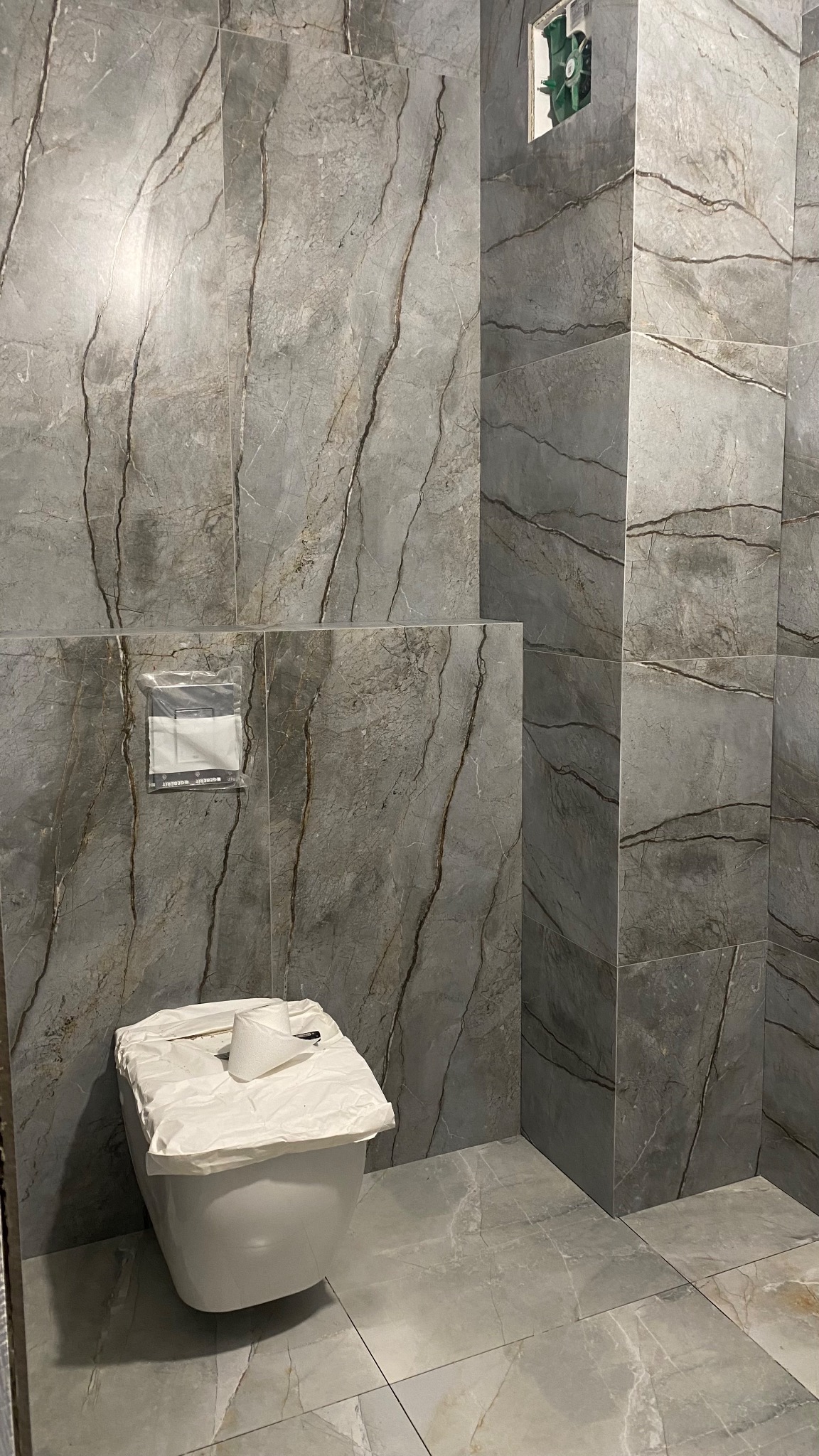 Nowa, podwieszana toaleta, zabezpieczona folią ochronną, w łazience wykończonej płytkami imitującymi marmur w odcieniach szarości z wyraźnym żyłowaniem. Widoczny fragment instalacji podtynkowych.