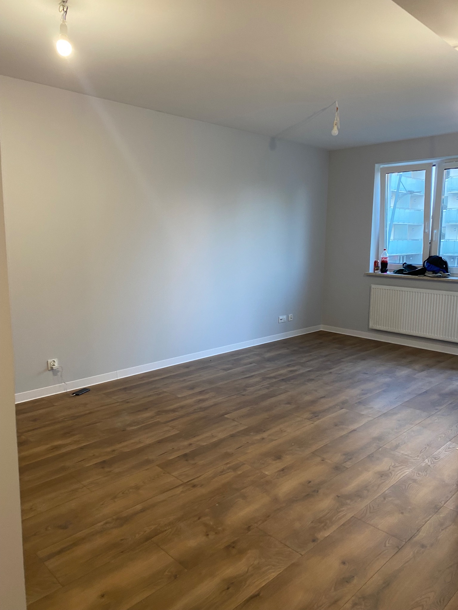 Pusty pokój po remoncie z ułożonymi panelami podłogowymi w odcieniach brązu, szare ściany, grzejnik pod oknem, widoczne gniazdka elektryczne i wiszące żarówki.
